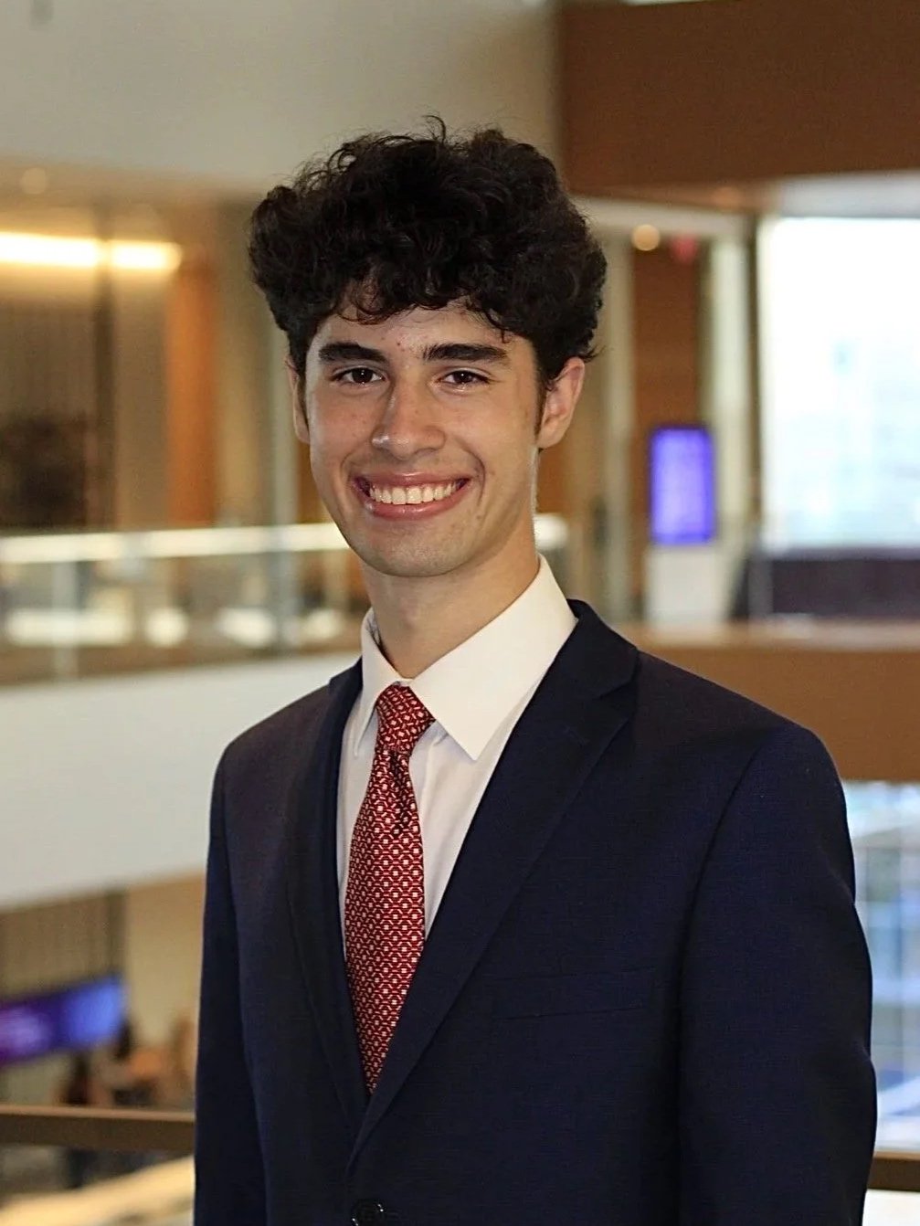 A young man with dark, curly hair, wearing a black suit, white shirt, and red patterned tie, smiling in an indoor setting.