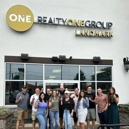 Group of people standing outside a real estate office with a sign that reads 'ONE Realty ONE Group Landmark.'