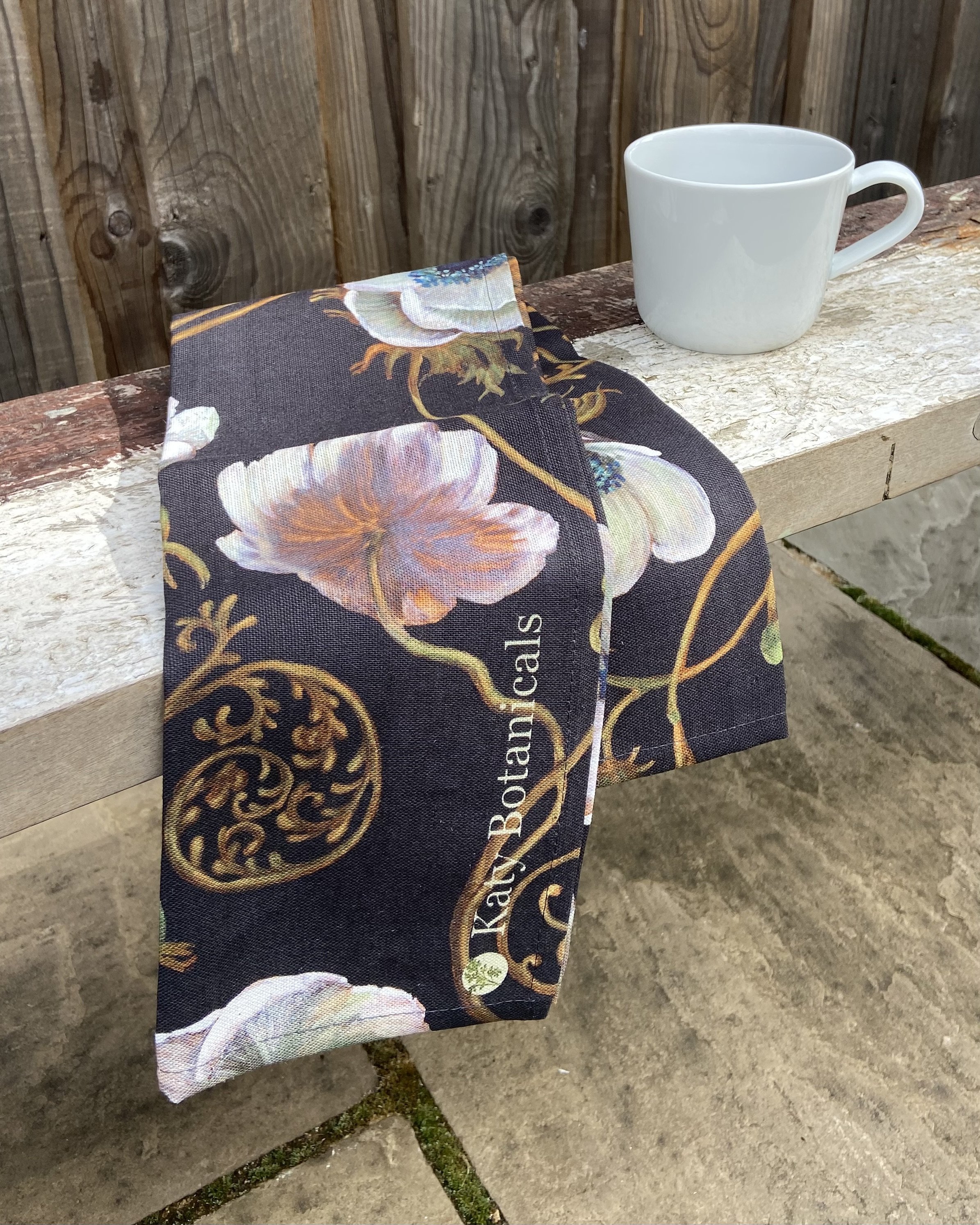 Photo of arts and crafts movement anemone and bird tea towel printed on heavy english linen, shown with a cup of tea on a work bench in front of wooden panelling