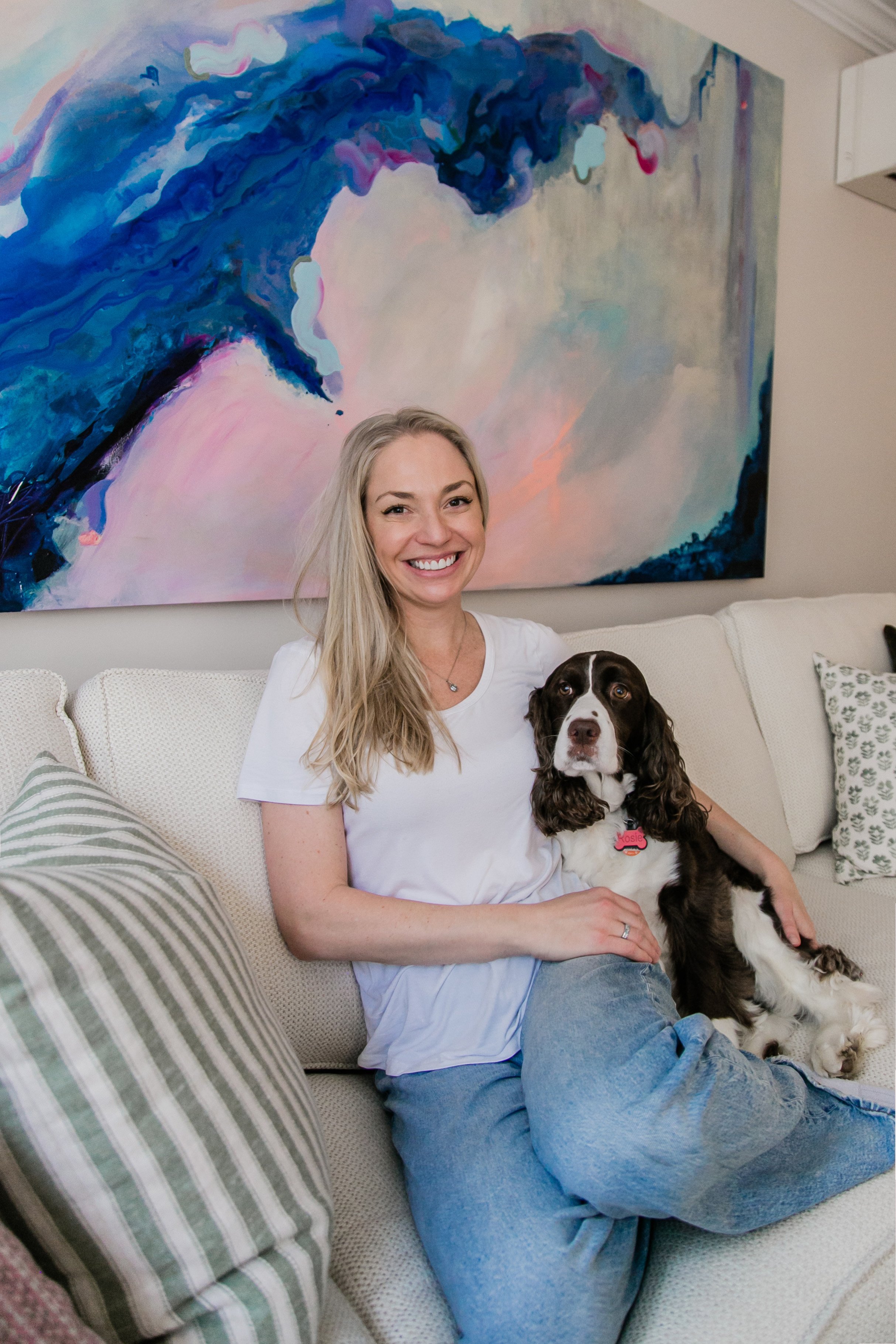 A woman with long blonde hair smiling and sitting on a cream-colored sofa with a brown and white dog. There is an abstract blue and pink painting on the wall behind them, and striped pillows on the sofa.