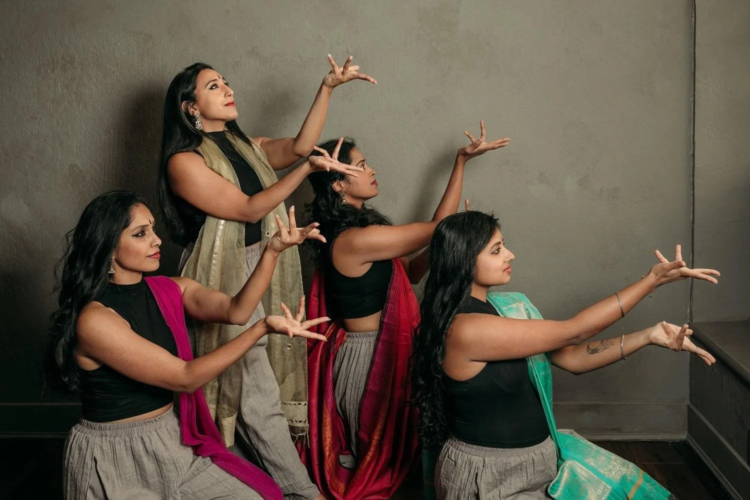 Five women in colorful traditional attire, sitting and standing against a plain wall, posing with intricate hand gestures.