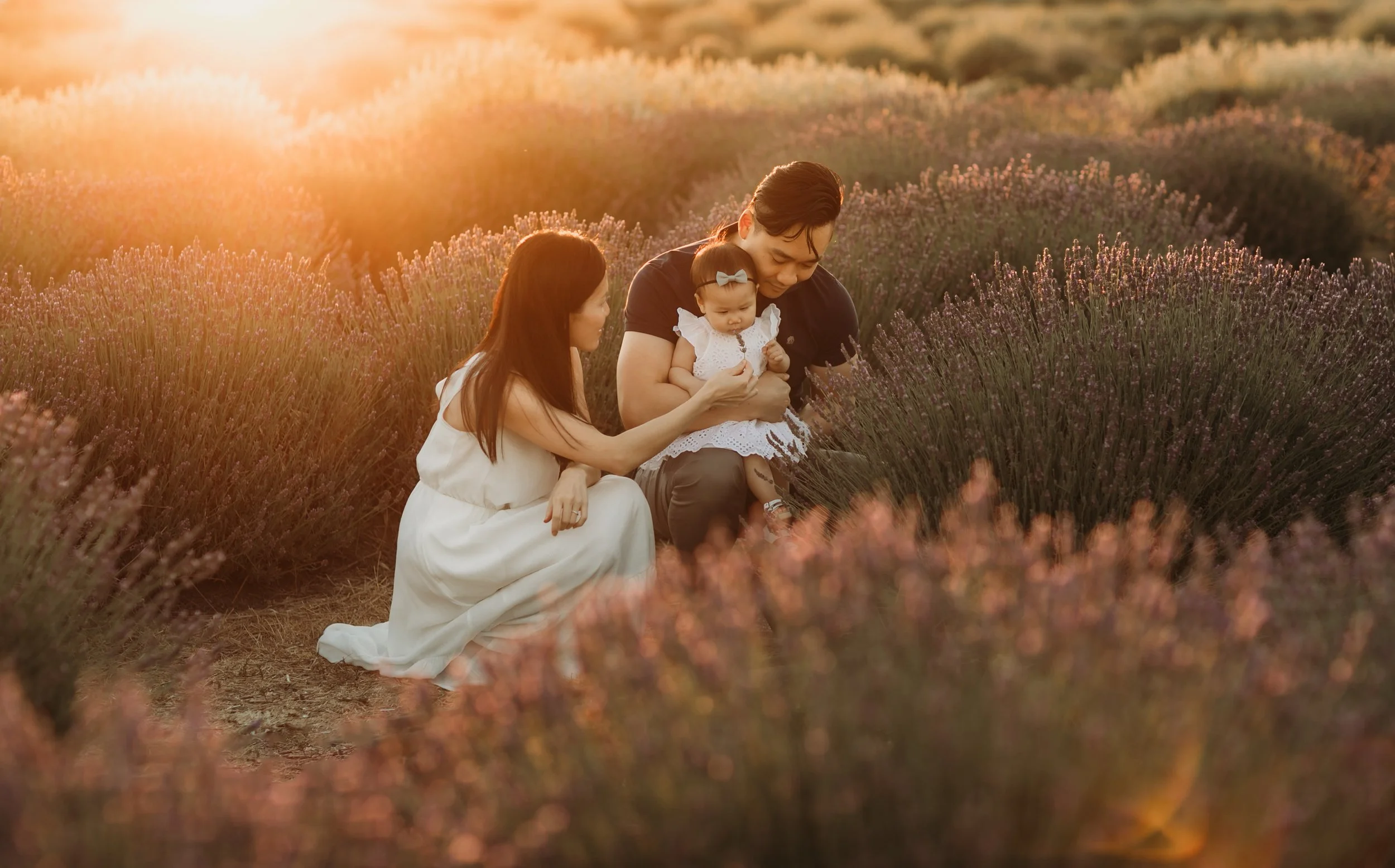 A family of three, a man, woman, and baby, sitting in a field of lavender during sunset, enjoying a moment together.