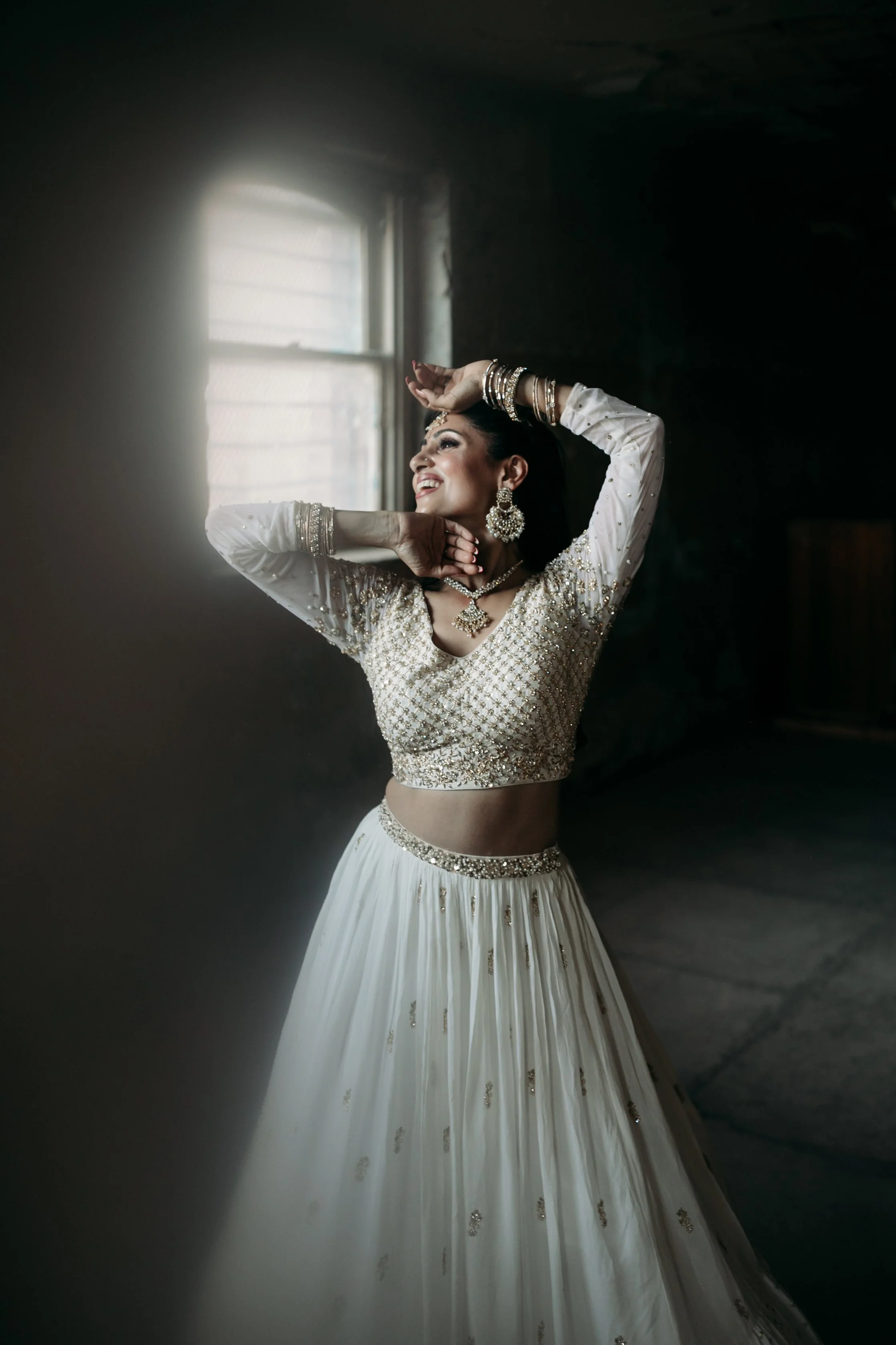 A woman in traditional Indian attire, jewelry, and makeup posing near a window inside a dark room.