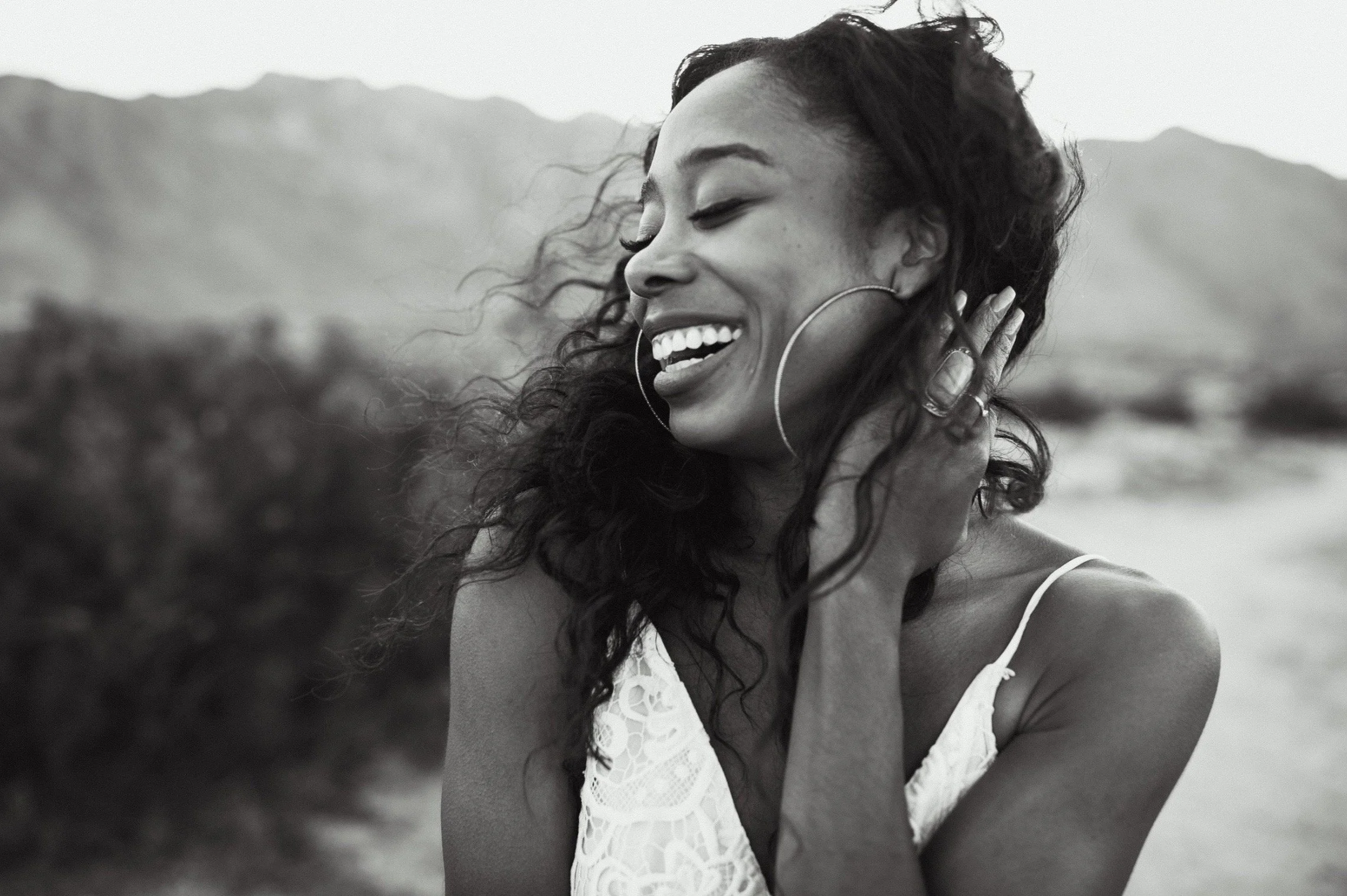 A black and white photo of a woman smiling with her eyes closed, holding her hand to her neck, outdoors with mountains in the background.