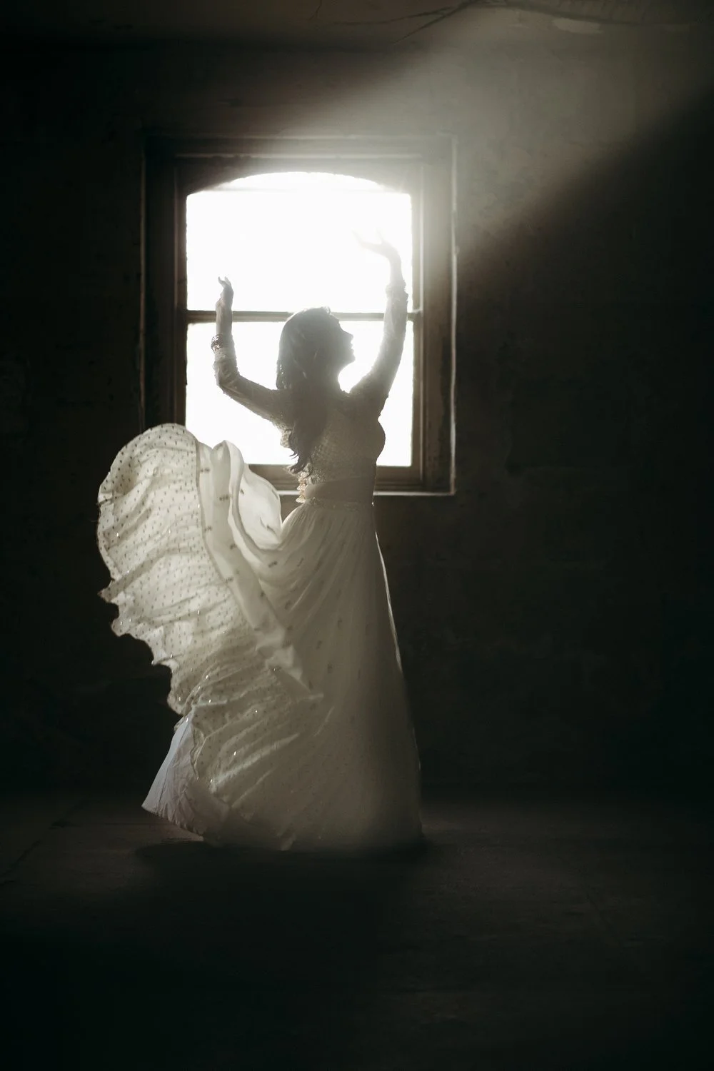 A woman in a flowing dress dancing or posing near a window with sunlight streaming in, creating a silhouette effect.