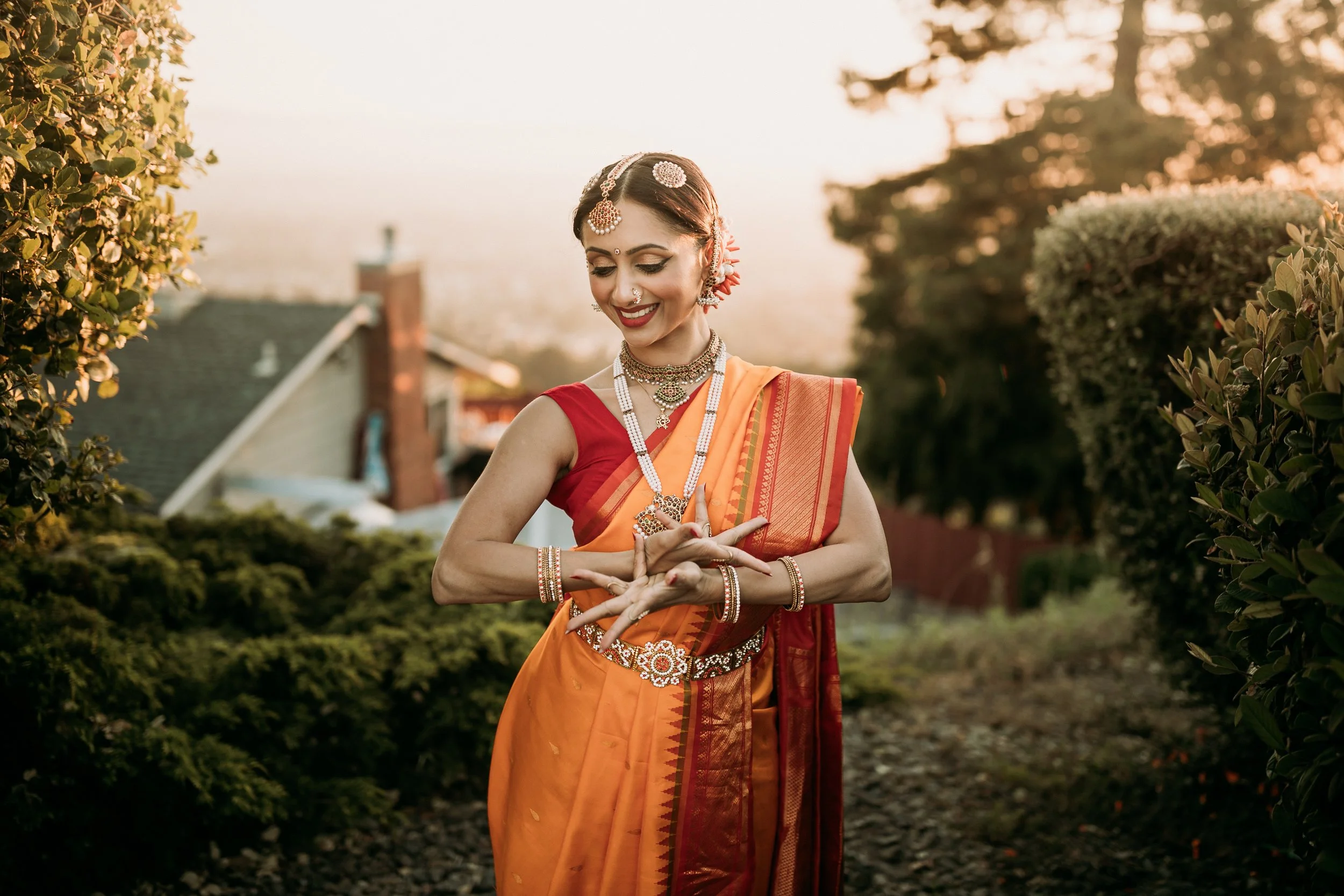 A woman dressed in traditional Indian attire, wearing an orange sari with red accents, is smiling and performing a dance outdoors during sunset.