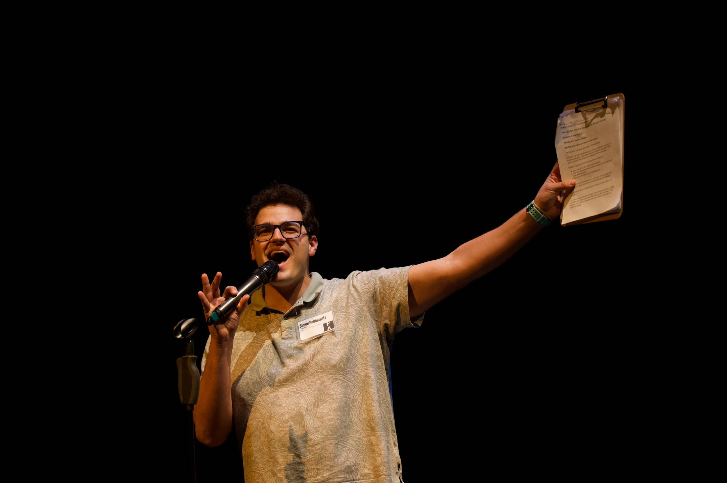 A young man with glasses speaking into a microphone on stage, holding up a clipboard with papers, wearing a beige patterned shirt, and a name badge reading "Simon Rabinowitz."