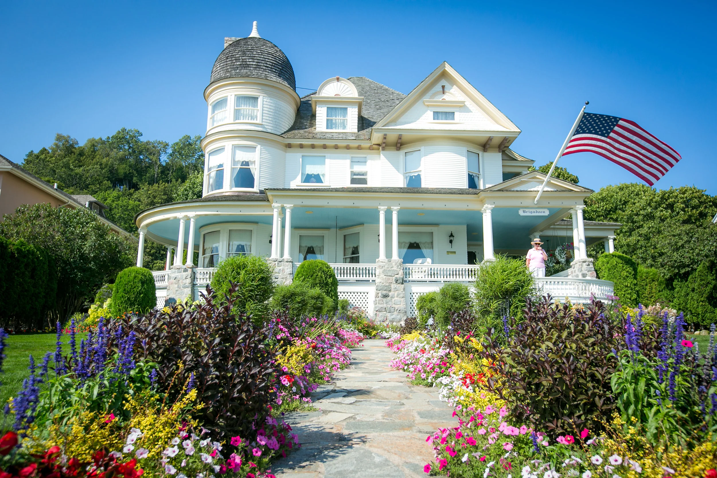 A large, white, Victorian-style house on Mackinac Island with a rounded turret and a porch, surrounded by vibrant flower beds and green bushes, with an American flag waving in the front yard under a clear blue sky.
