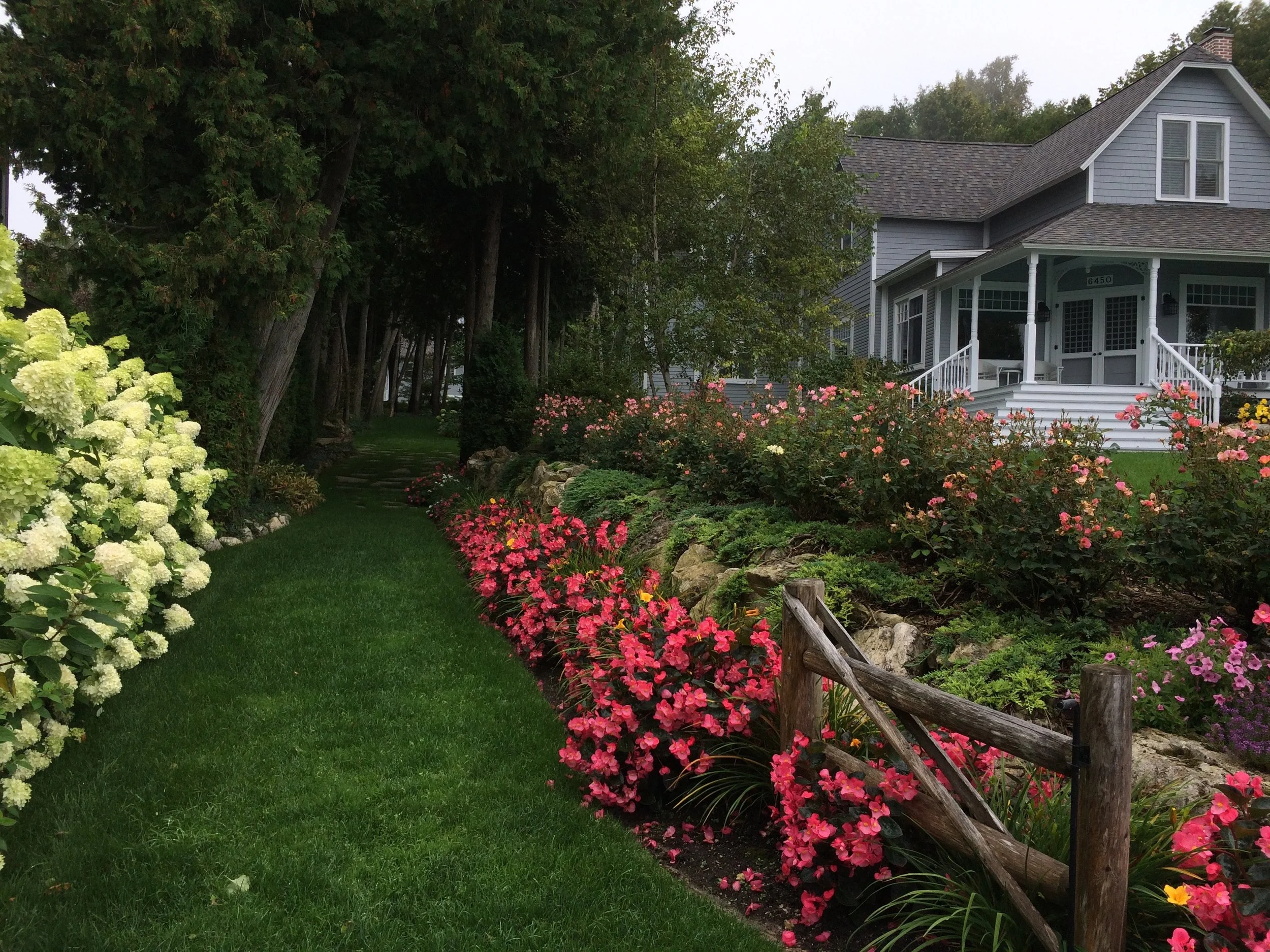 A gray house with a porch surrounded by a lush garden with pink, white, yellow, and purple flowers, and green trees and grass.