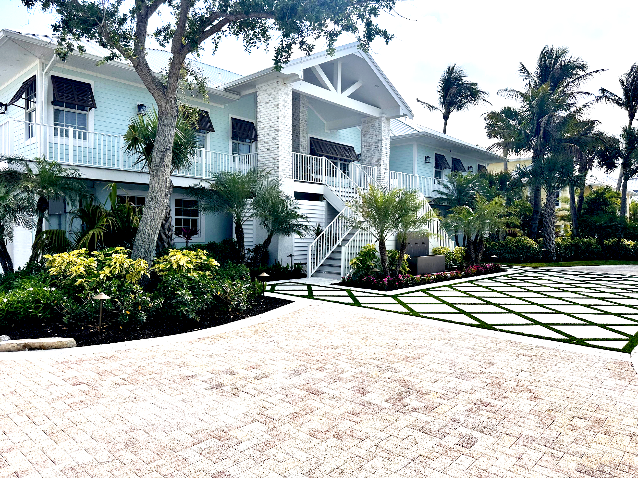 A light blue two-story house with white accents, surrounded by palm trees and lush greenery, with a paved walkway and a garden area in the foreground.