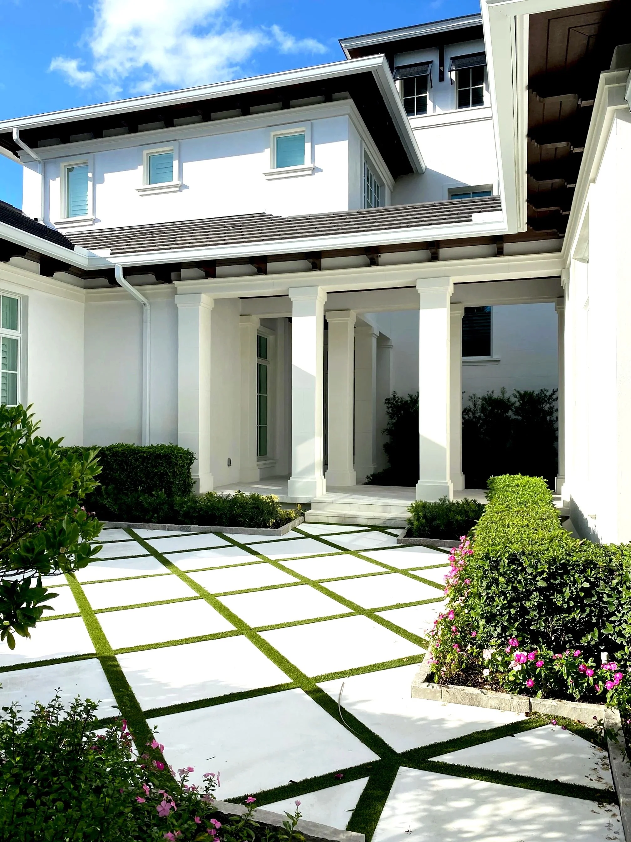 White house with a well-maintained front yard featuring a geometric pattern of white paving stones and green grass, surrounded by lush bushes and pink flowers.