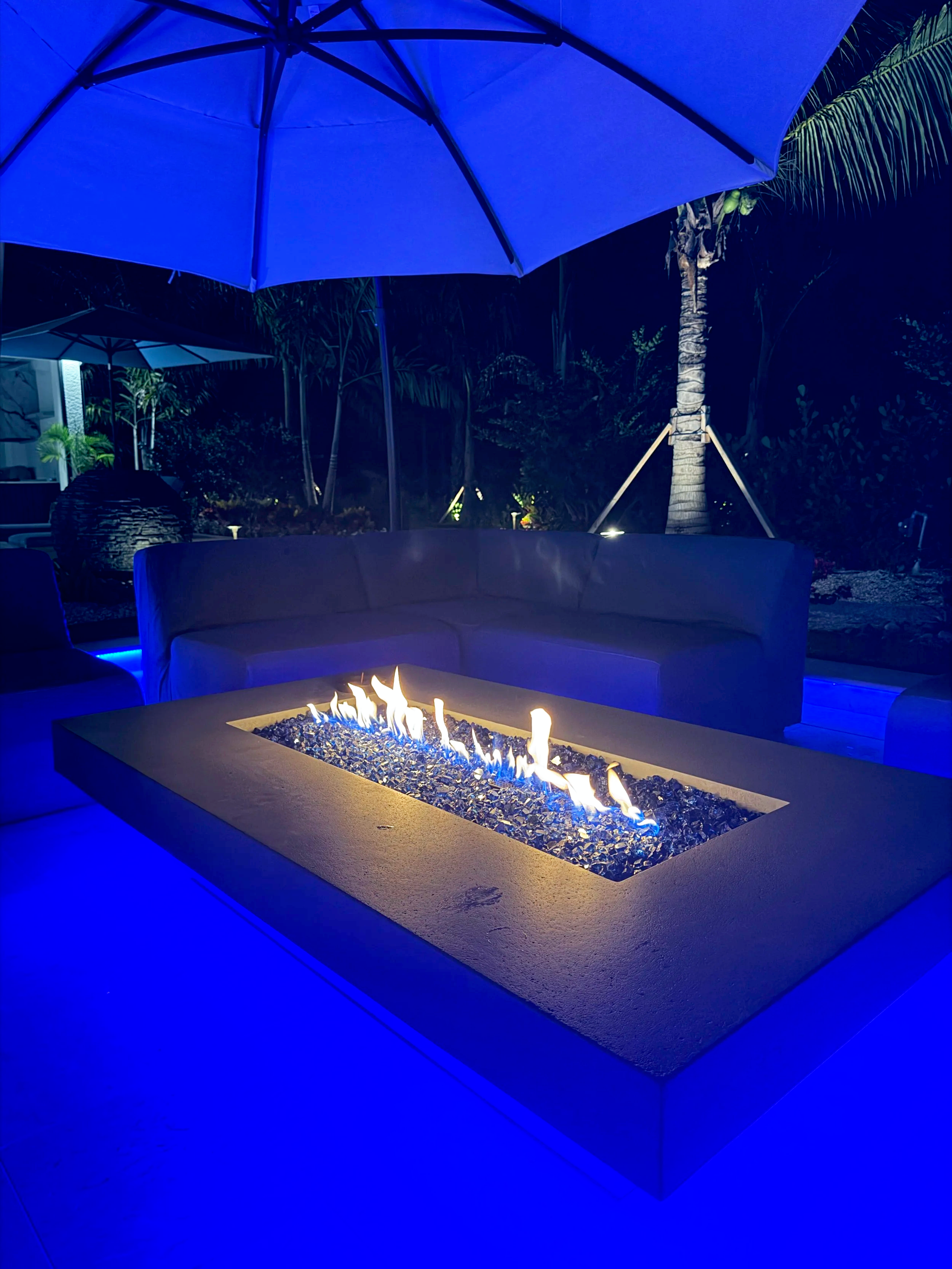 Outdoor patio area at night with a custom rectangular fire pit, a round outdoor sofa, large patio umbrellas, and lush green tropical plants including a palm tree illuminated by ambient lighting.