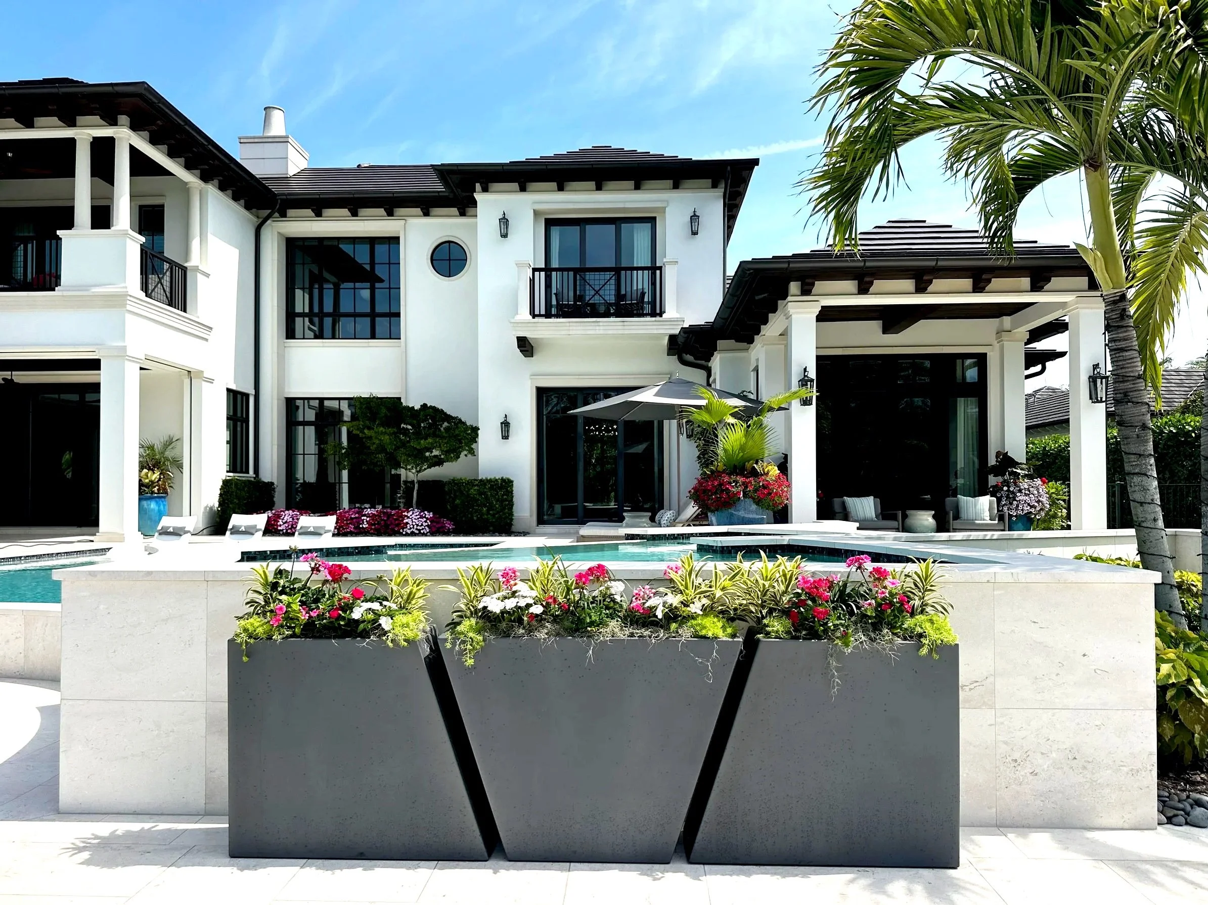 A modern white house with black accents, surrounded by lush greenery, including a tall palm tree on the right side. In the foreground, a large gray planter with colorful flowers sits in front of a swimming pool, and an outdoor seating area with chair