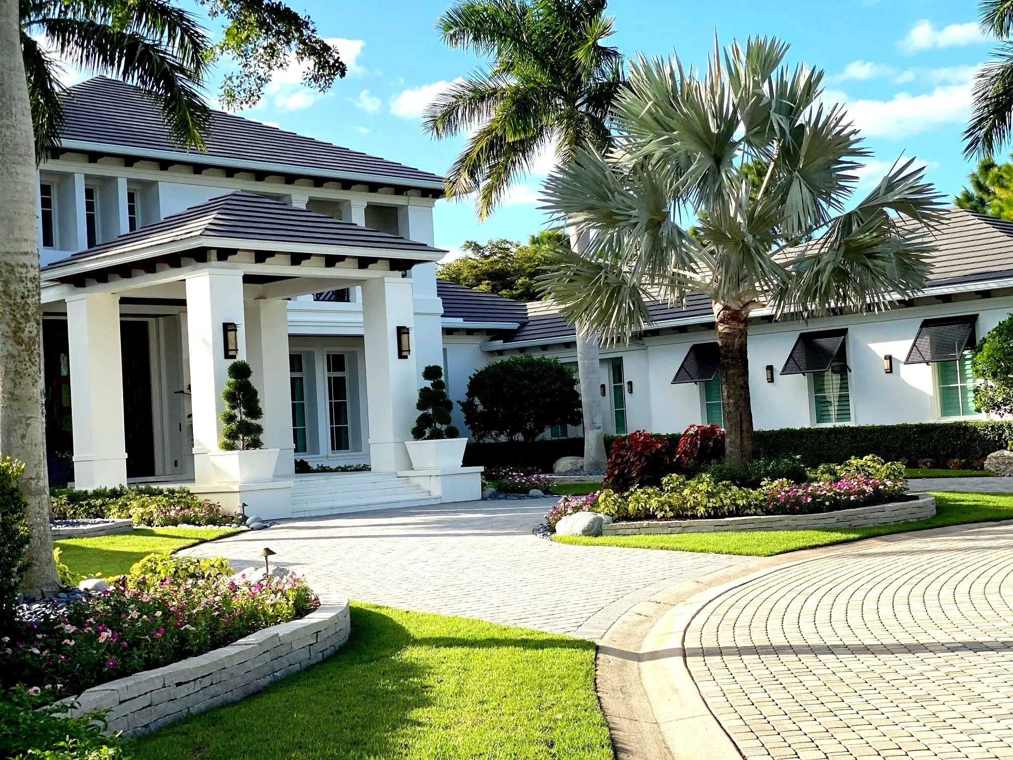 A modern white house with a gray tiled roof, surrounded by well-maintained landscaped gardens, including a curved driveway, flowering plants, and palm trees under a blue sky.