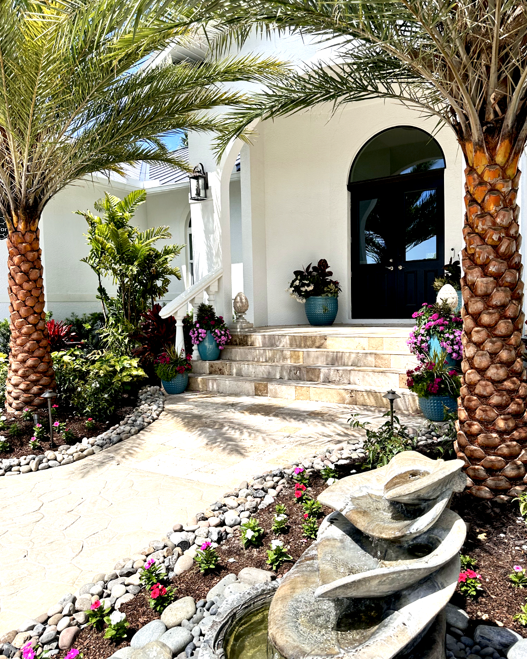 Front entrance of a Southern Florida white house with black door, stone steps, surrounded by tropical plants, pink flowers, two palm trees, and a stone water fountain in the garden.