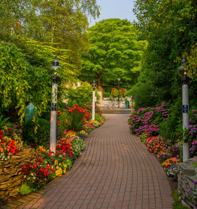 A brick pathway in a lush garden surrounded by colorful flowers and trees, with vintage-style lampposts lining the path.