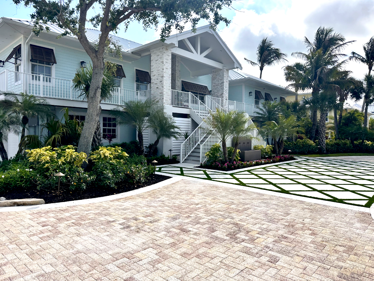 A light blue two-story house with white trim, a staircase, and a balcony, surrounded by tropical plants and palm trees, with a paved driveway in the foreground and a cloudy sky overhead.