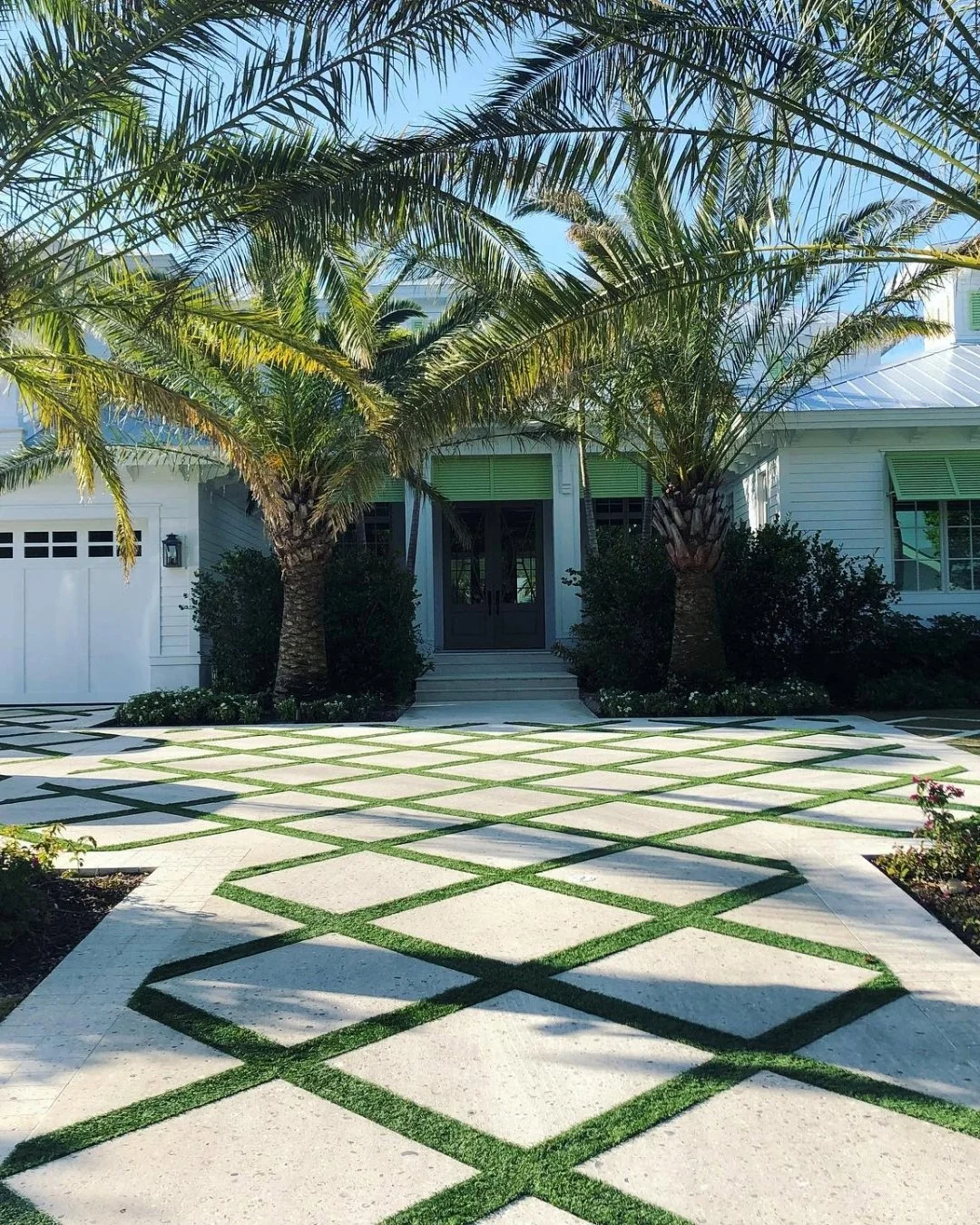 Front yard of a white house with two palm trees, a patterned walkway with grass in between, and bushes near the entrance.