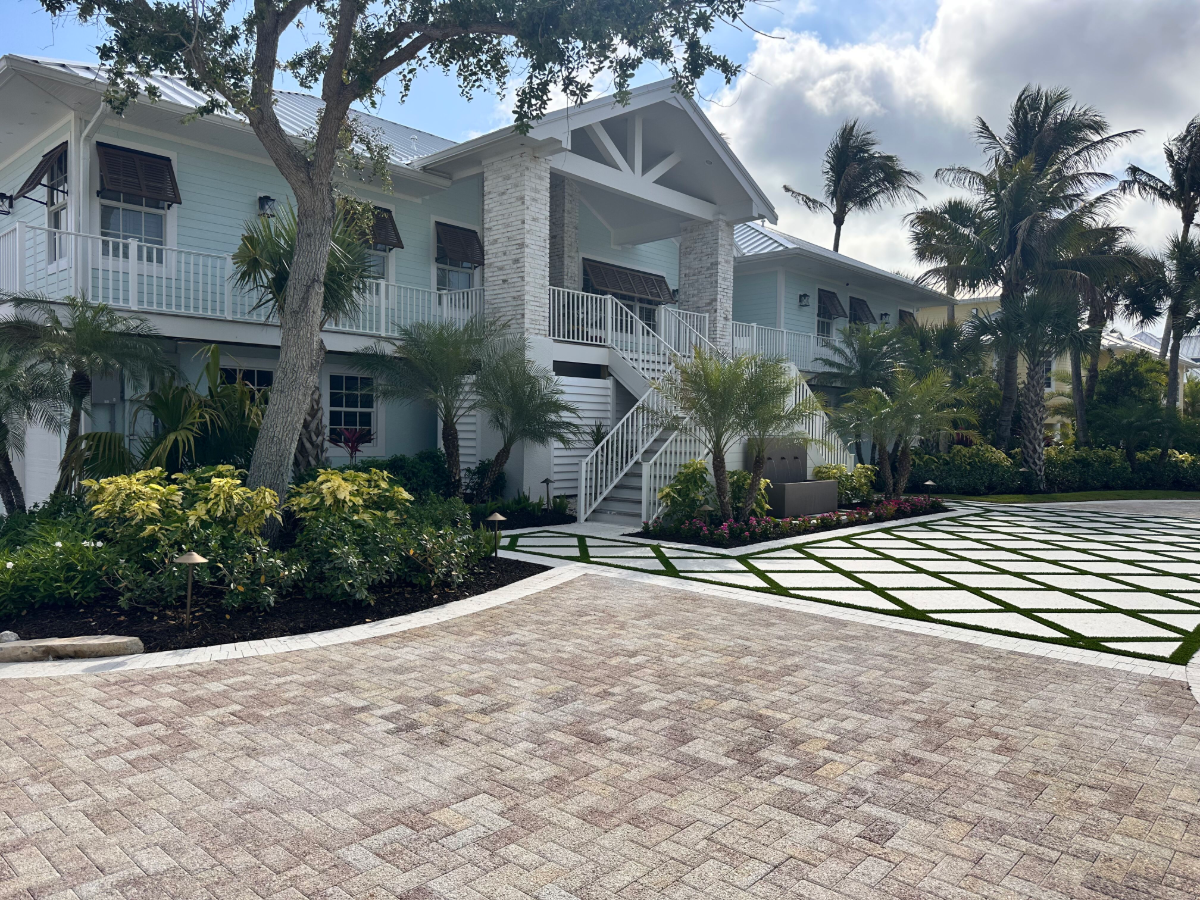 A light blue house with a white railing and stairs leading up to the second floor, surrounded by palm trees and lush greenery, with a patterned driveway in the foreground.