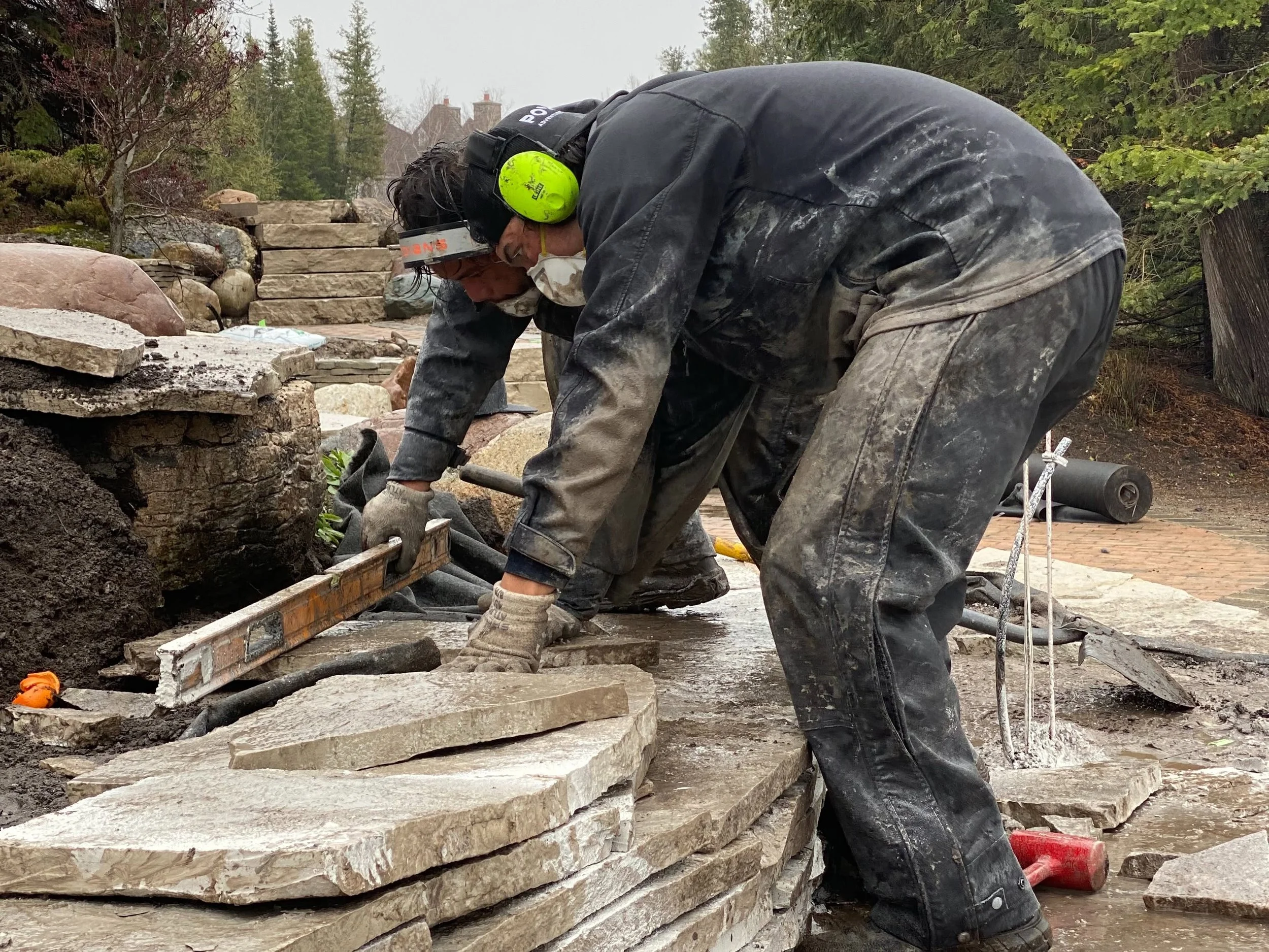 Landscape construction crew members wearing a black jackets and muddy pants working on building a stone wall outdoors, surrounded by trees and greenery, with a cloudy sky overhead.