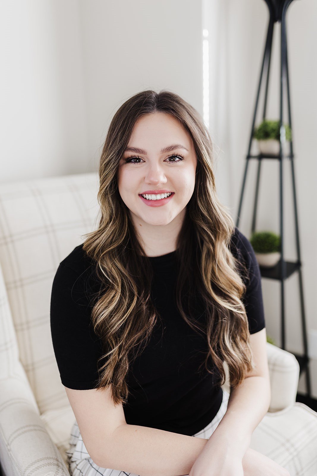 A young woman with long wavy brown hair, smiling, wearing a black top, sitting on a beige plaid couch in a bright room with a black metal plant stand with small potted plants in the background.