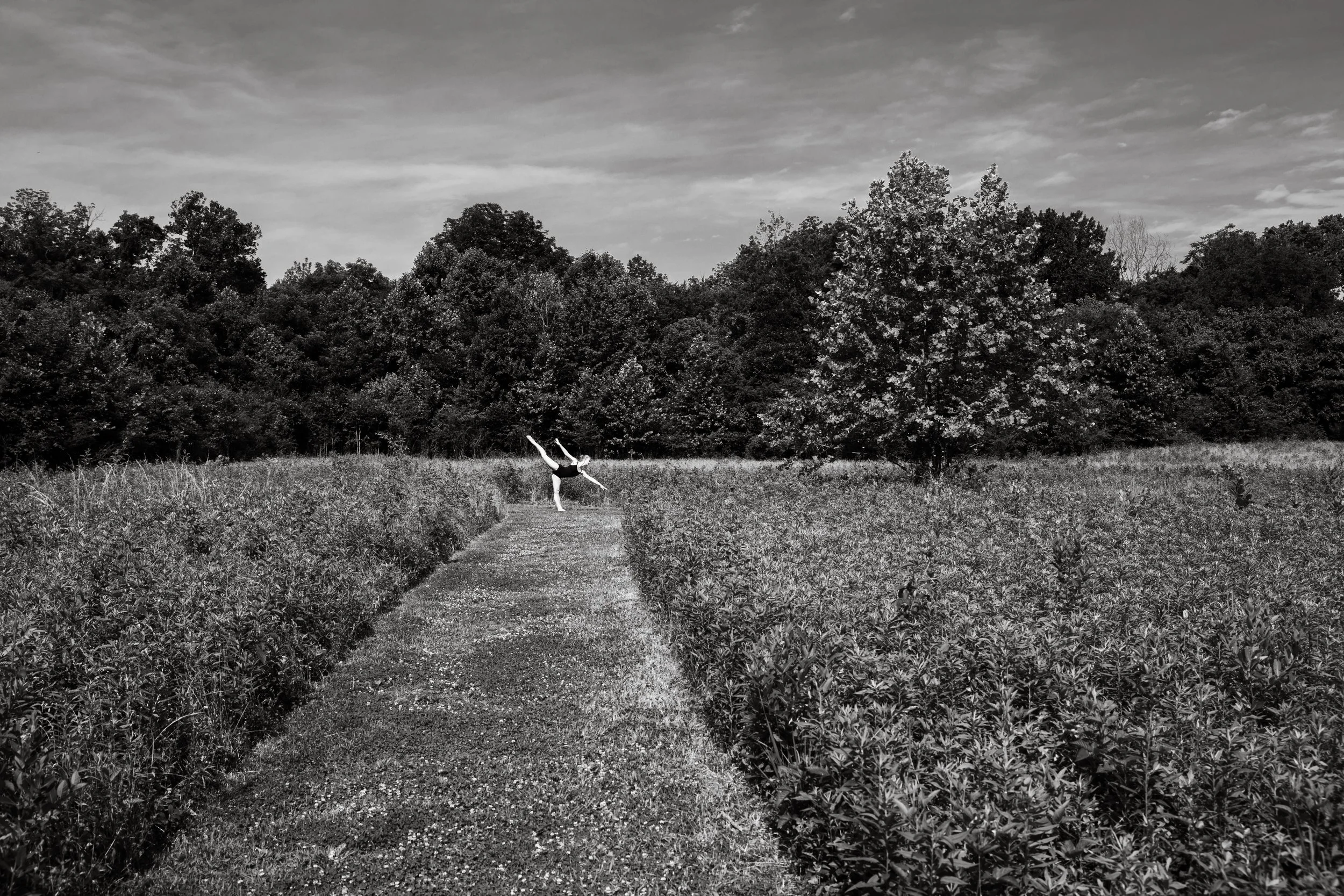 A person in a dance pose standing on one leg with arms extended, on a narrow dirt path in a field with tall grass or crops, surrounded by trees with a cloudy sky in the background, in black and white.
