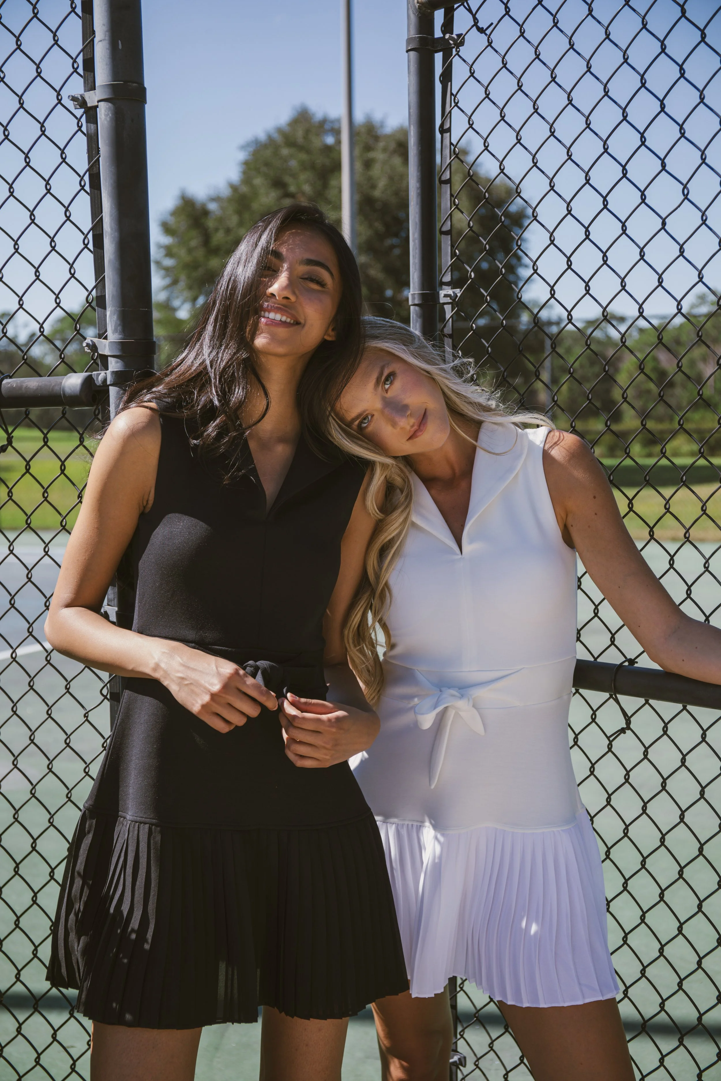 Two women standing close together in front of a chain-link fence, smiling at the camera on a sunny day. One woman has long dark hair and is wearing a black sleeveless dress, and the other has long blonde hair and is wearing a white sleeveless dress.