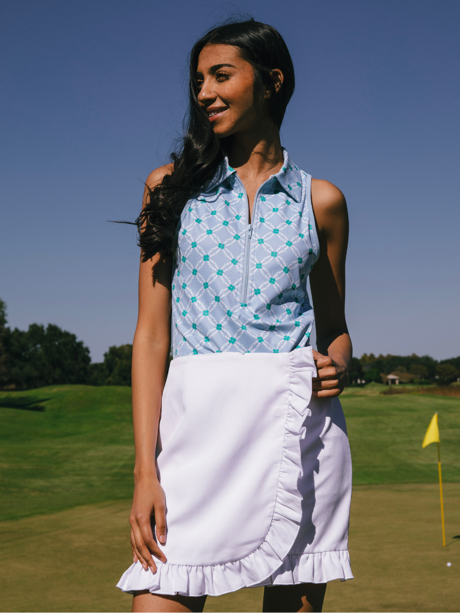 A woman with long dark hair smiling on a golf course, wearing a sleeveless blue patterned top and a white ruffled skirt near a yellow flag.