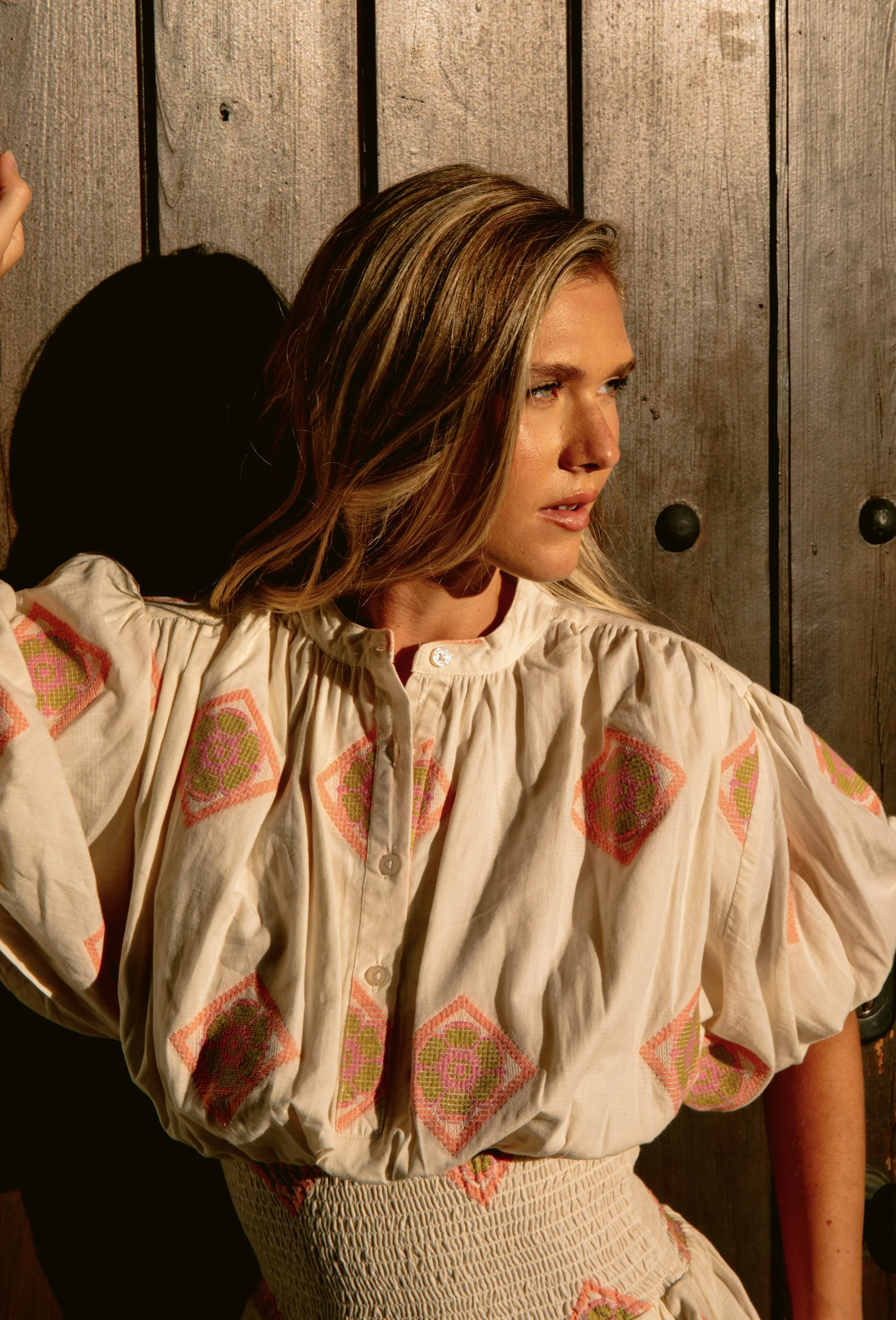 A woman with blonde hair and wearing a cream-colored blouse with embroidered pink and green patterns, posing against a wooden backdrop.