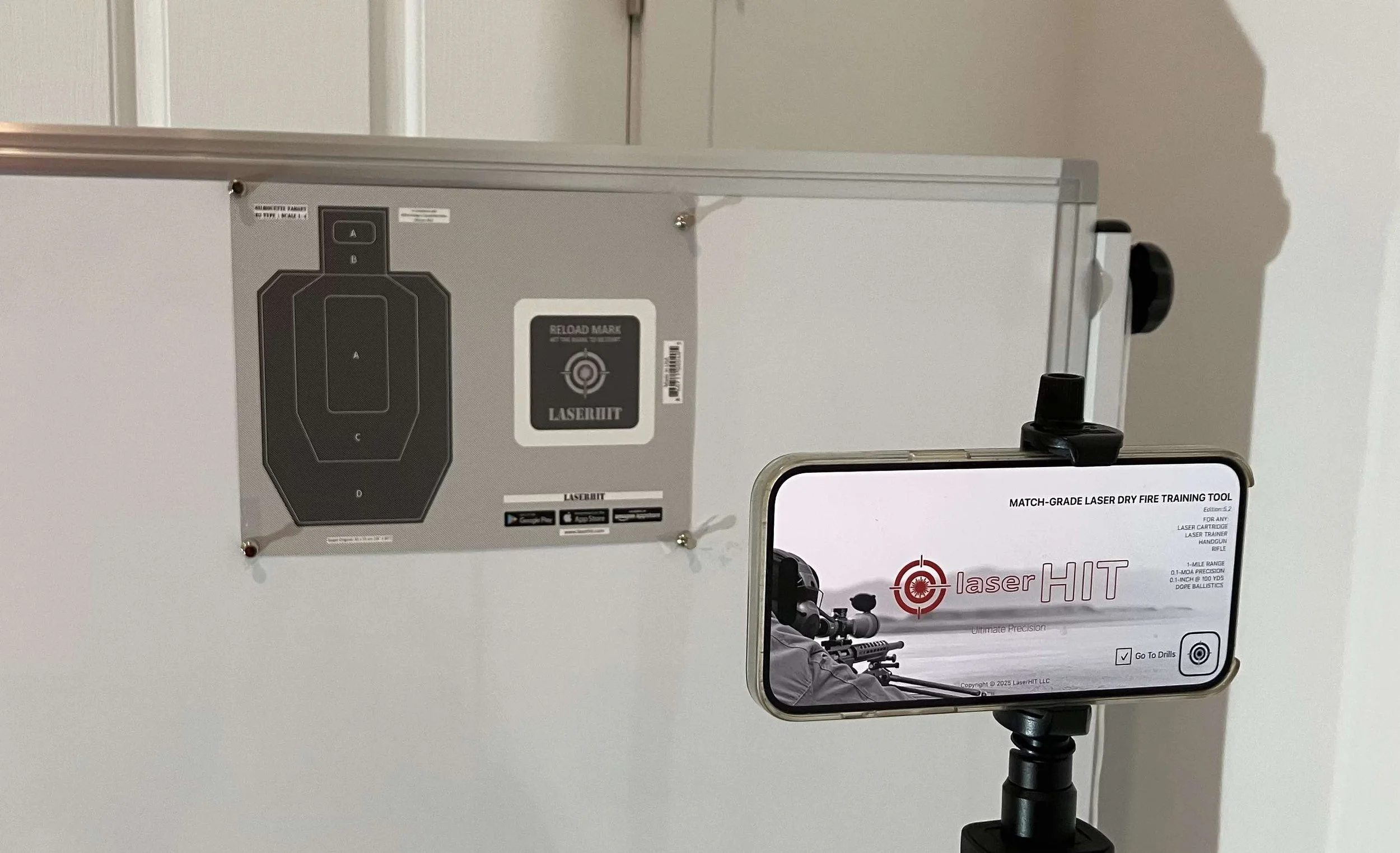 LaserHIT DRILLS training setup with a printed target on the wall and a smartphone positioned on a tripod, using the rear camera to detect laser hits during dry-fire practice