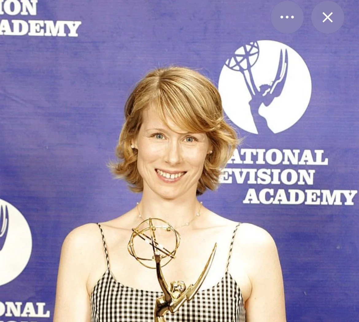 Woman with short red hair holding an Emmy award, standing in front of a blue backdrop with the text 'National Television Academy' and a logo.