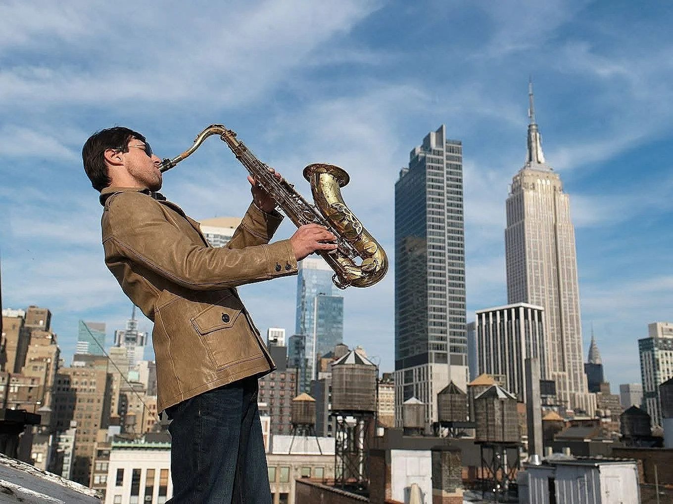 A man wearing sunglasses and a tan jacket playing a saxophone on a rooftop, with the New York City skyline, including the Empire State Building, in the background.