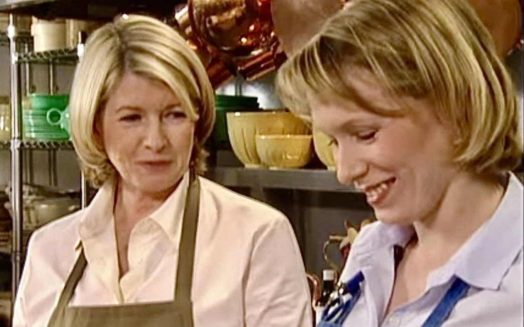 Two women in a kitchen, one with blonde hair wearing a white shirt and apron, the other also with blonde hair wearing a white shirt with blue apron, preparing food.