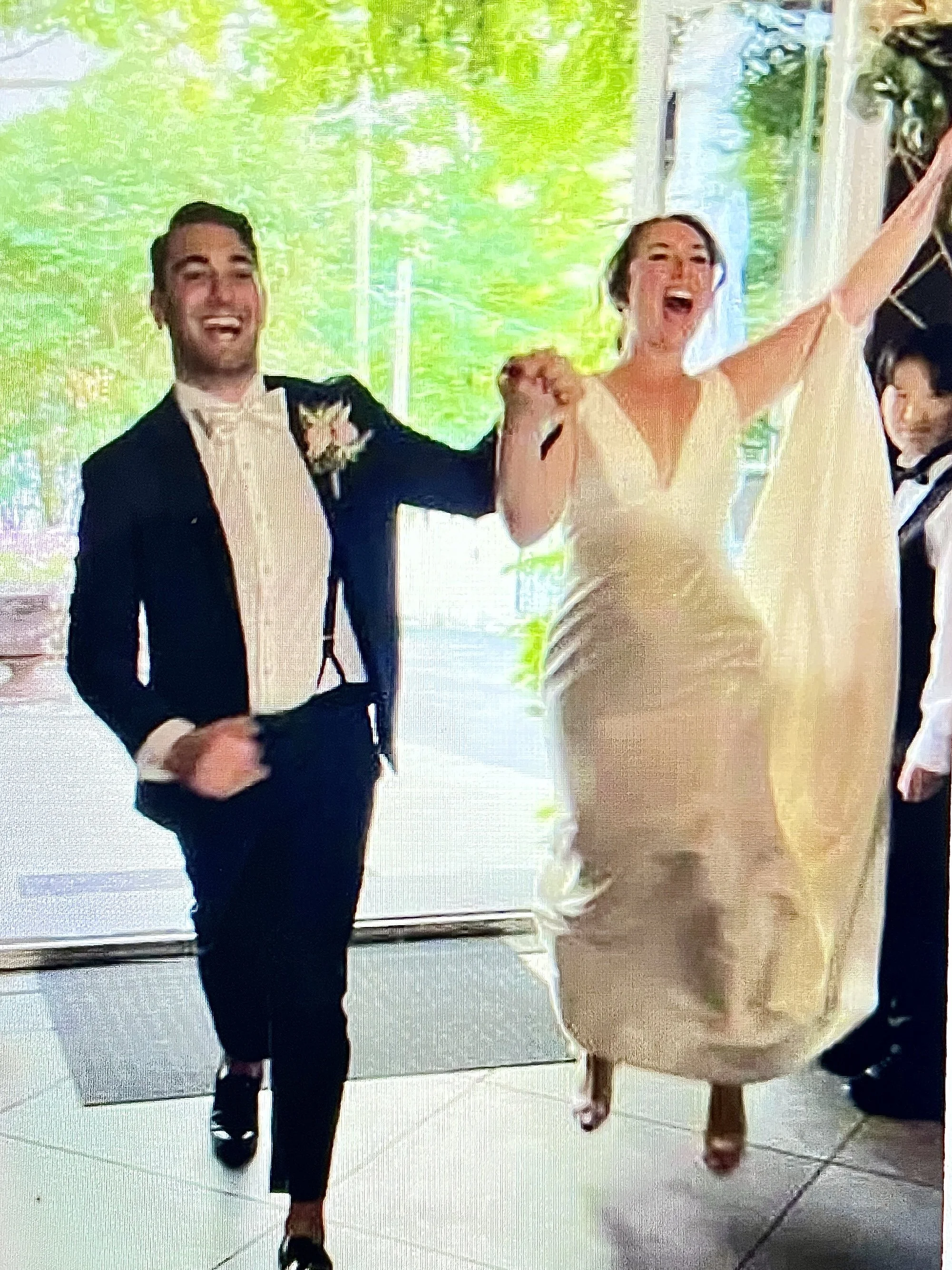 The bride and groom are joyfully entering together indoors, smiling and holding hands. They appear happy and celebratory.