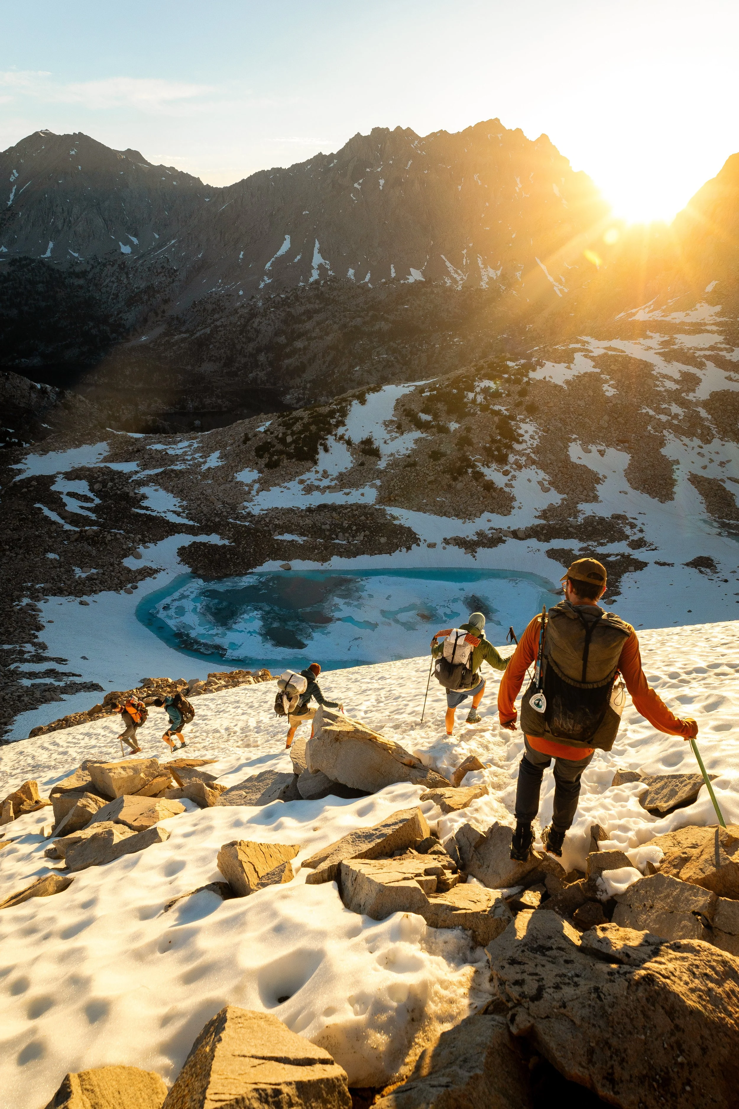 Mountaineers climbing snowy slope in mountainous terrain during sunset, with rugged rocks in foreground and a mountain lake in the background.