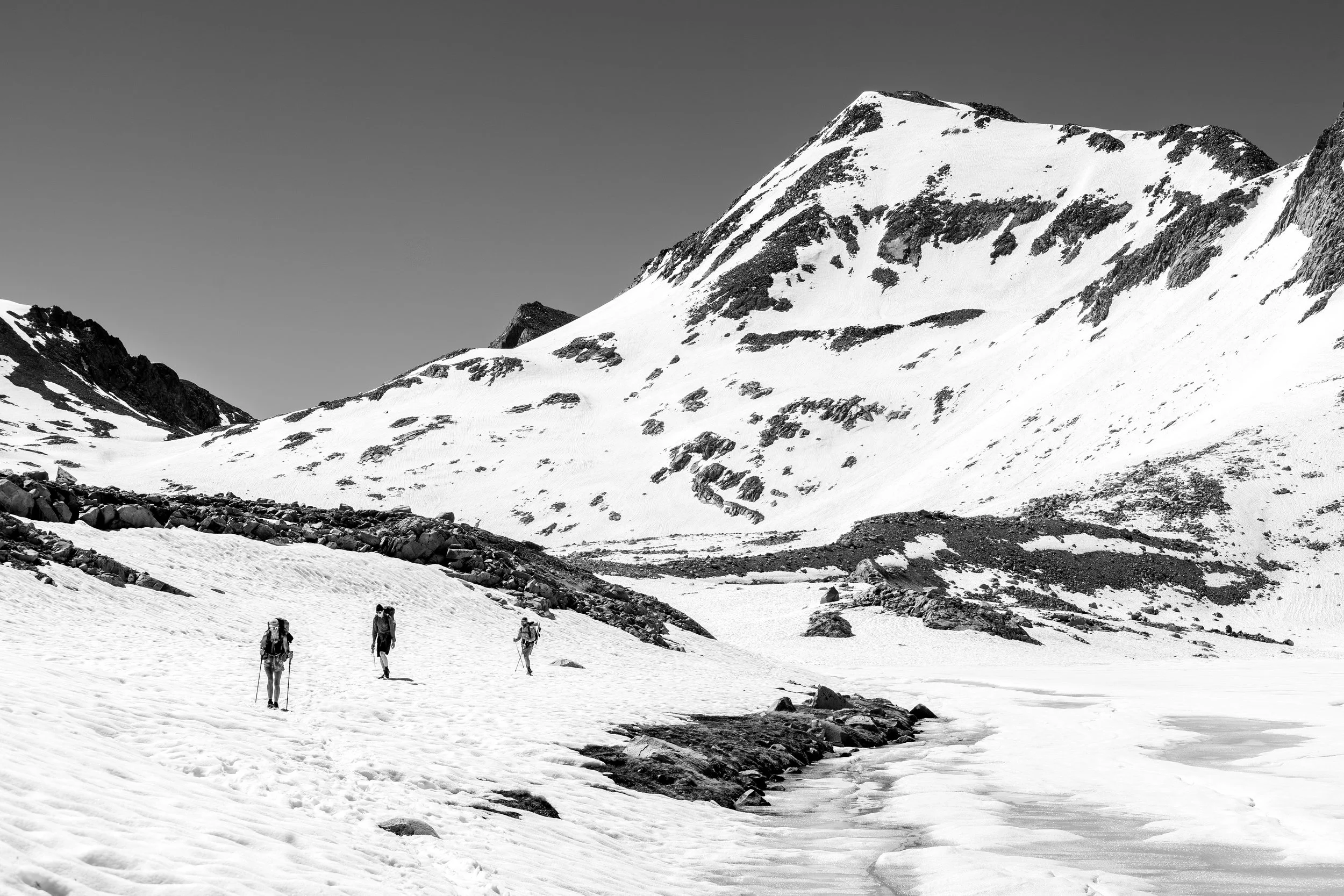 Four hikers with backpacks walking on a snowy mountain landscape with peaks in the background in black and white.