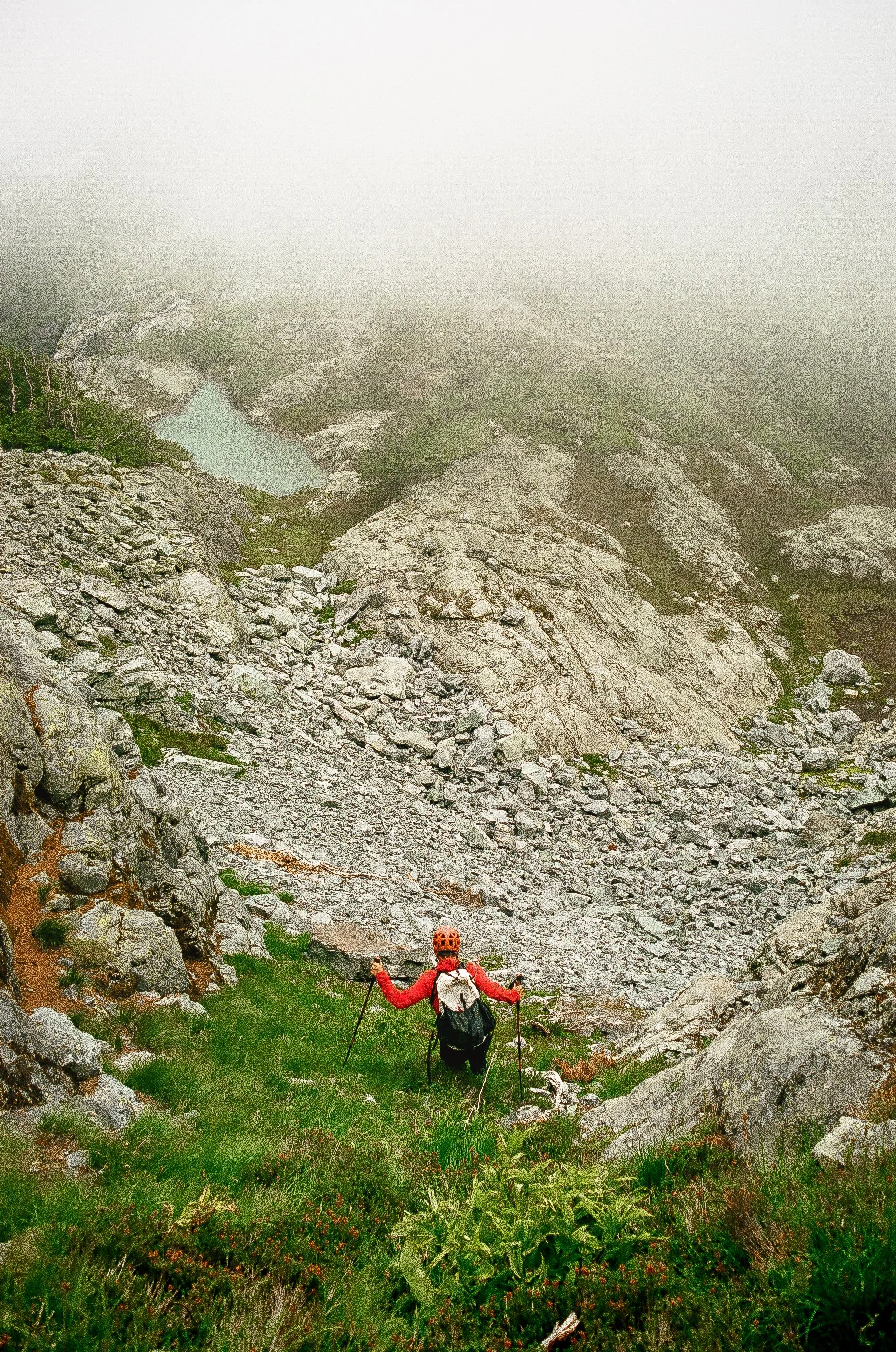 A mountain climber wearing an orange helmet and red jacket with trekking poles approaching a foggy, rocky mountain landscape with a small lake.