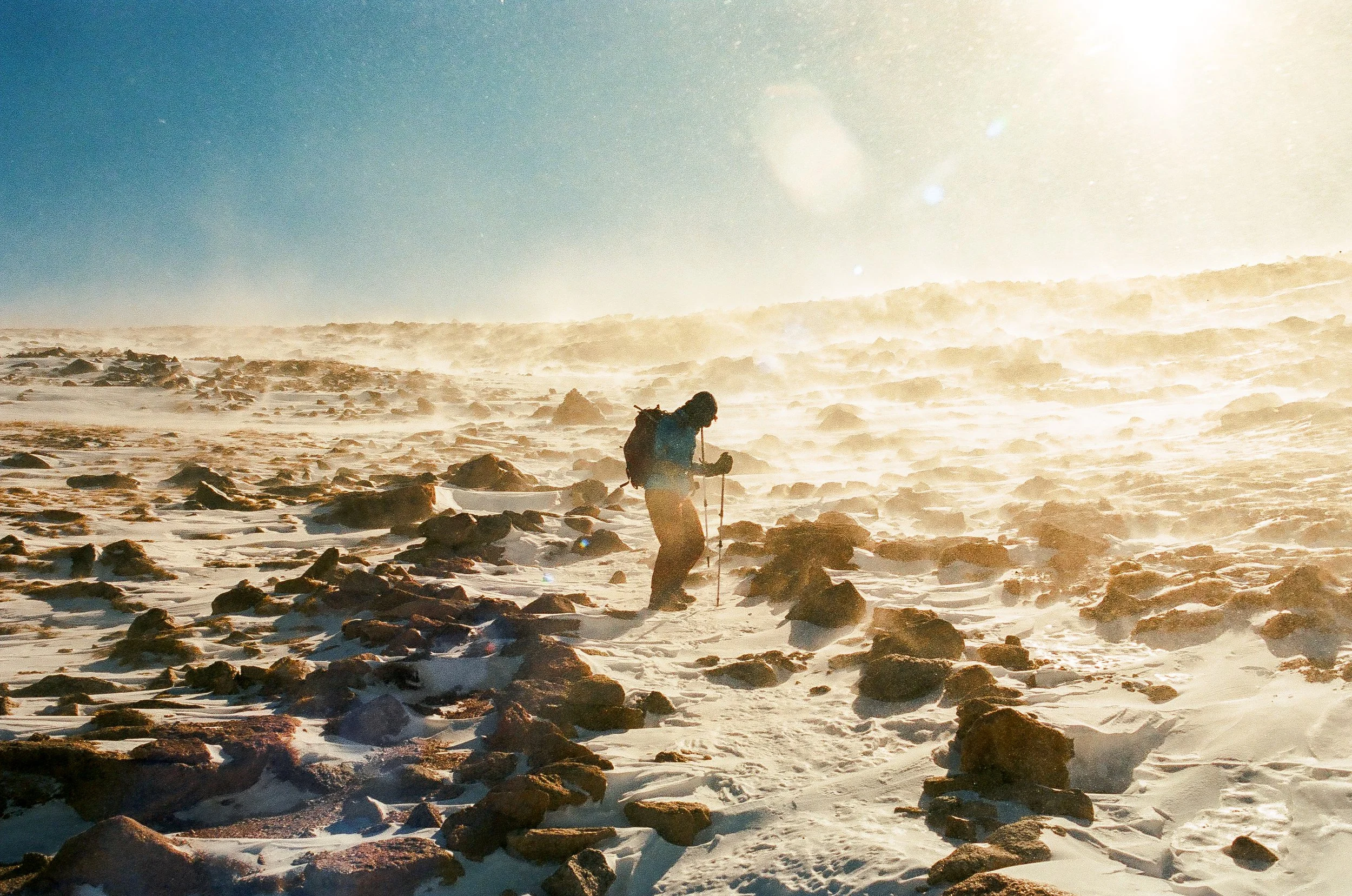 A person hiking through a snow-covered, rocky landscape during sunset or sunrise, with sunlight creating a bright glow in the sky.