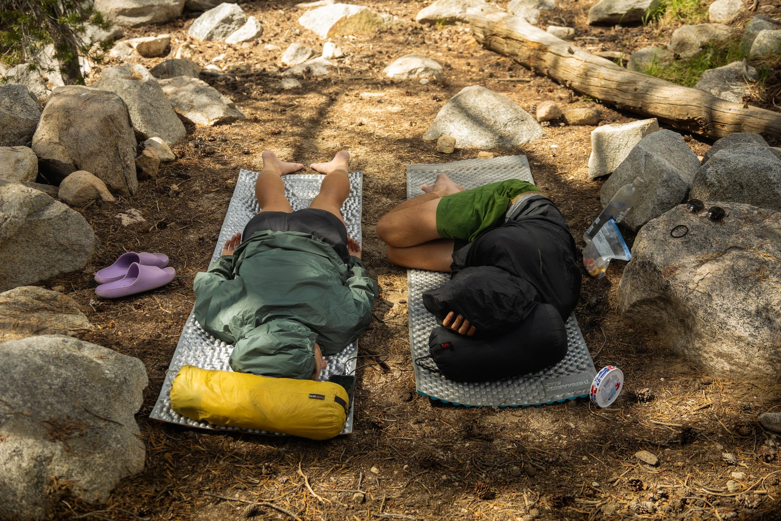 Two people lying on camping mats in a wooded outdoor area surrounded by rocks and trees, with personal items like shoes, a water bottle, sunglasses, and a container nearby.