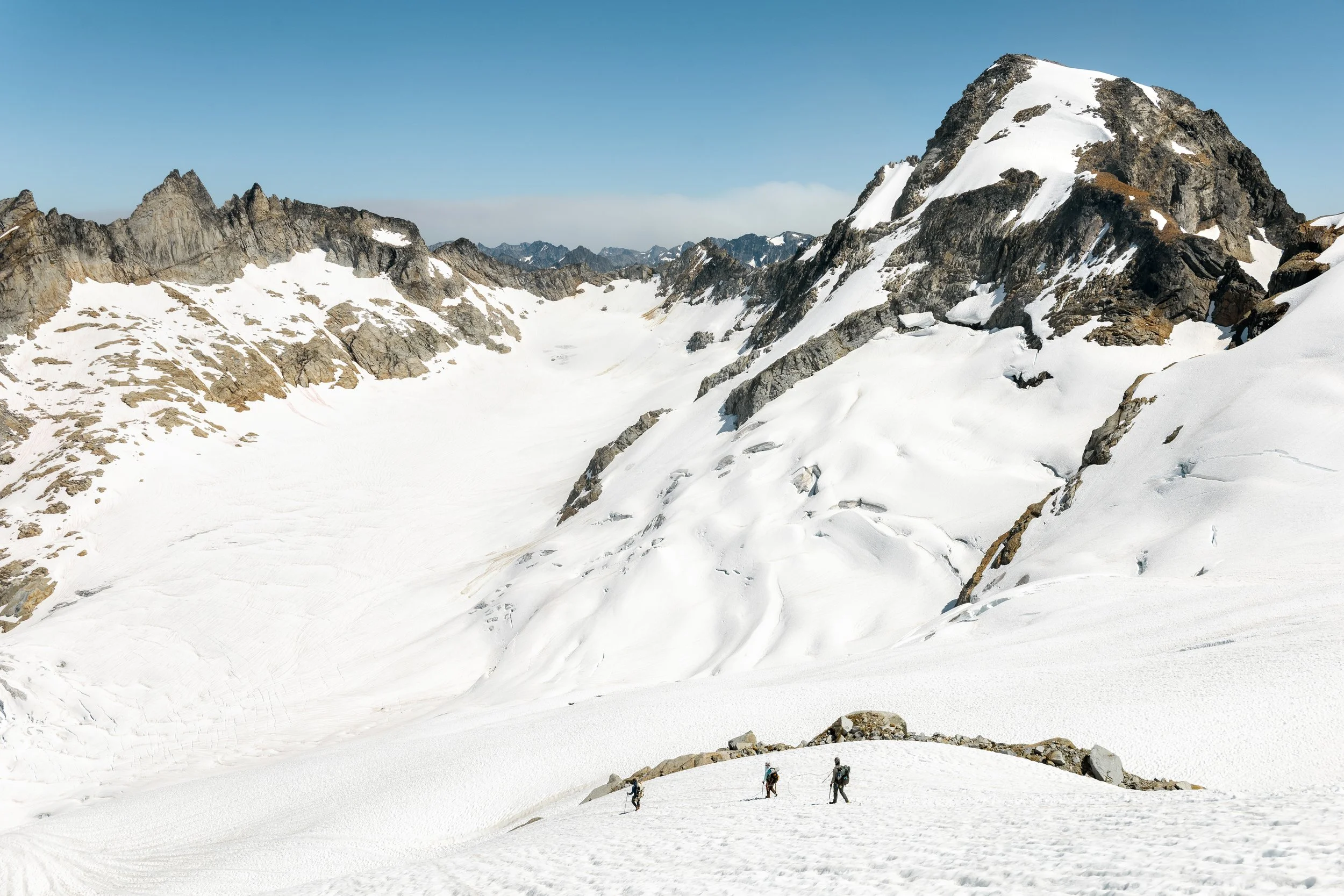 A group of four hikers walking across a snow-covered mountain landscape, with steep rocky peaks in the background under a clear blue sky.