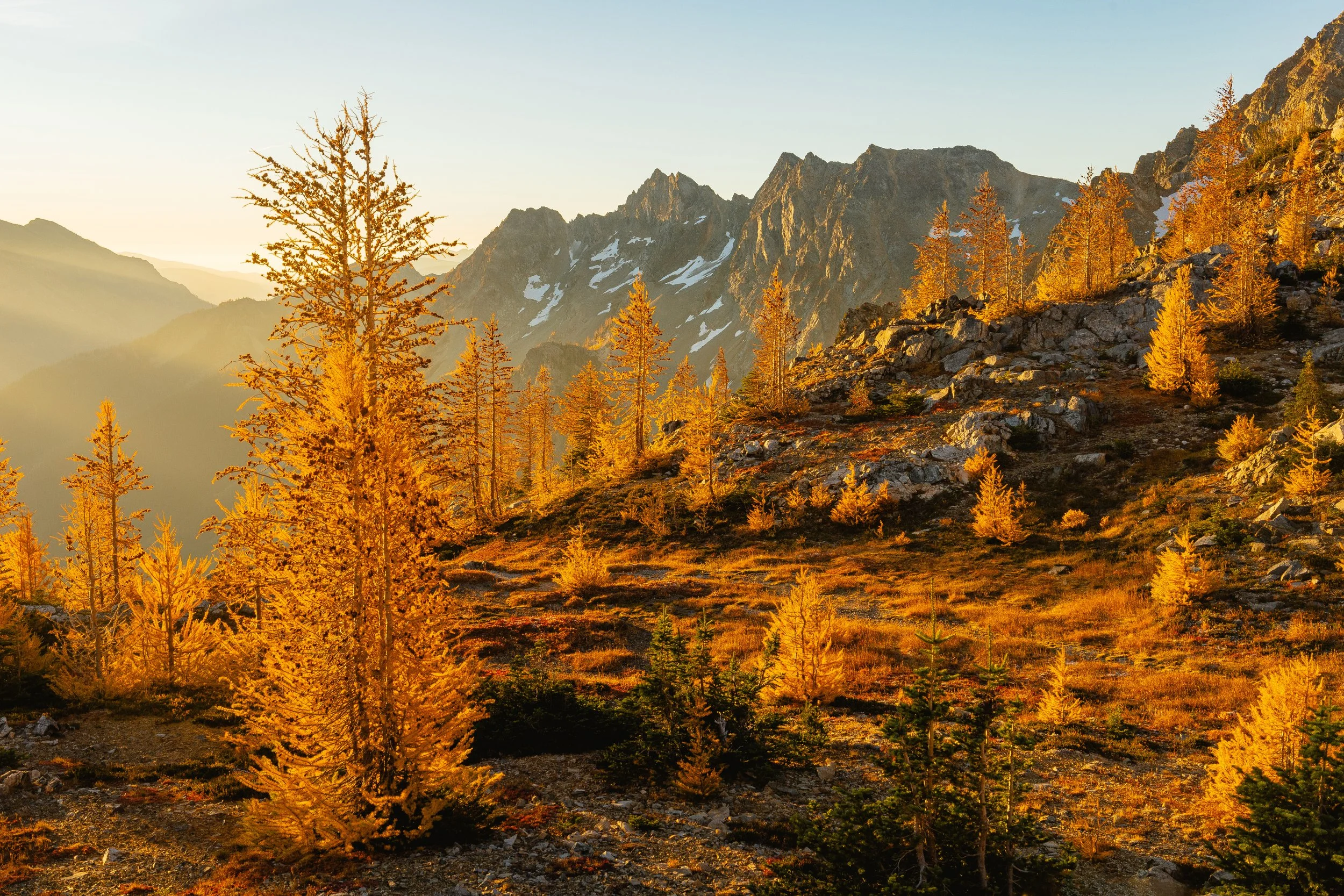 Sunlit mountain landscape with orange and yellow trees in the foreground and rugged peaks with patches of snow in the background.