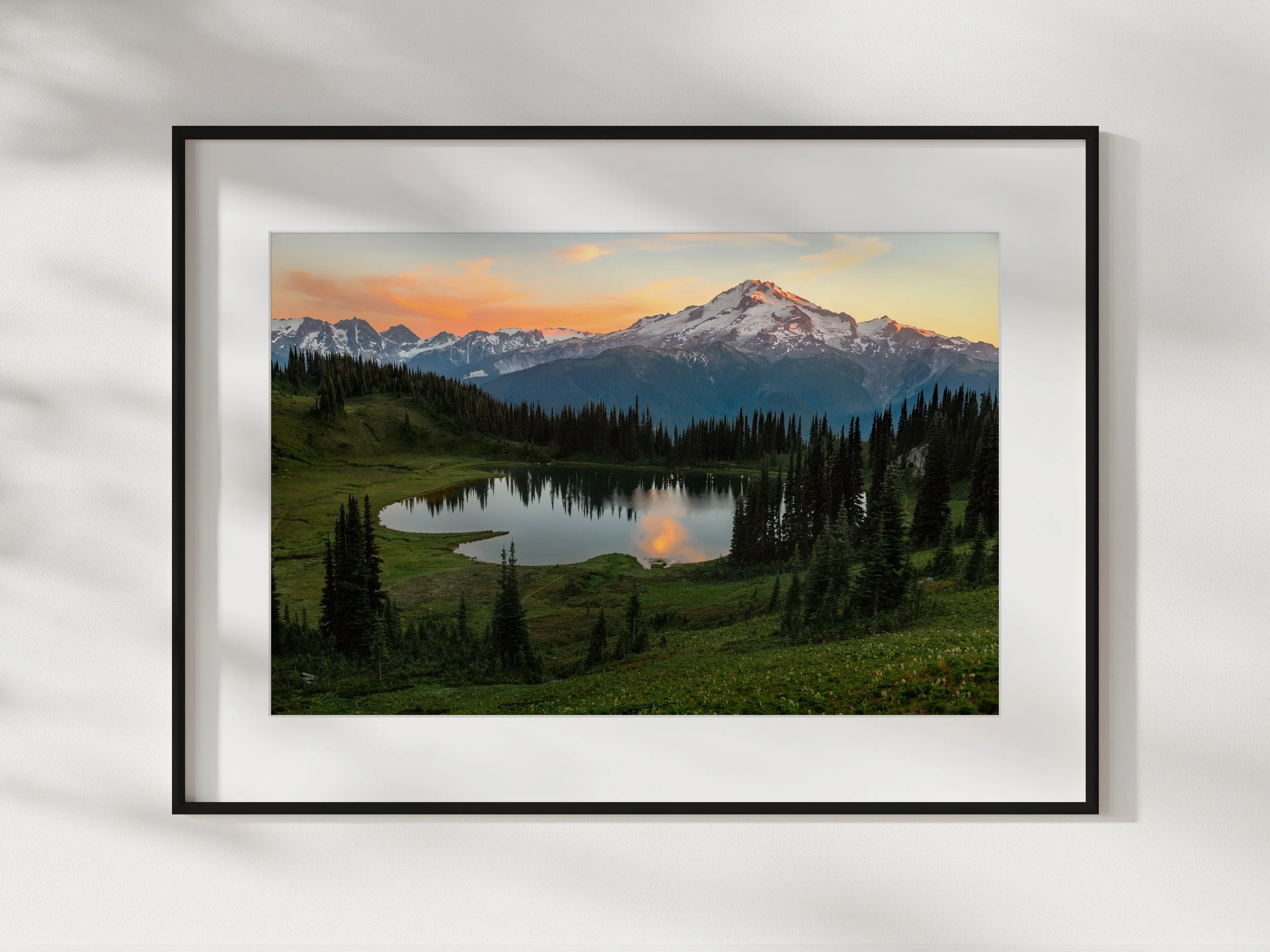Framed landscape photograph of snow-capped mountains, green hills, a forest, and a small lake during sunset.