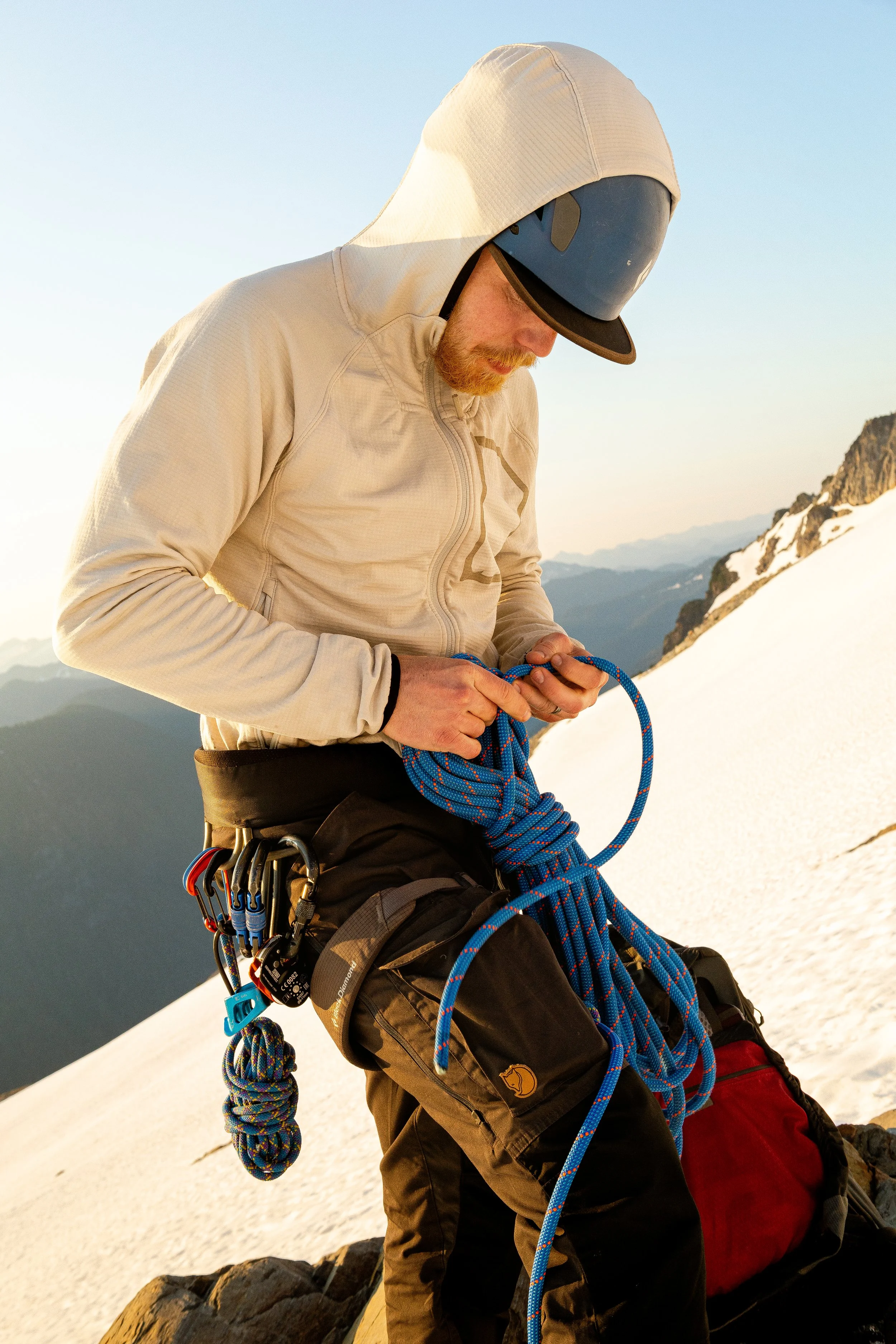 A man wearing a beige hoodie, black cap, and climbing gear adjusting a blue climbing rope on snowy mountain terrain.