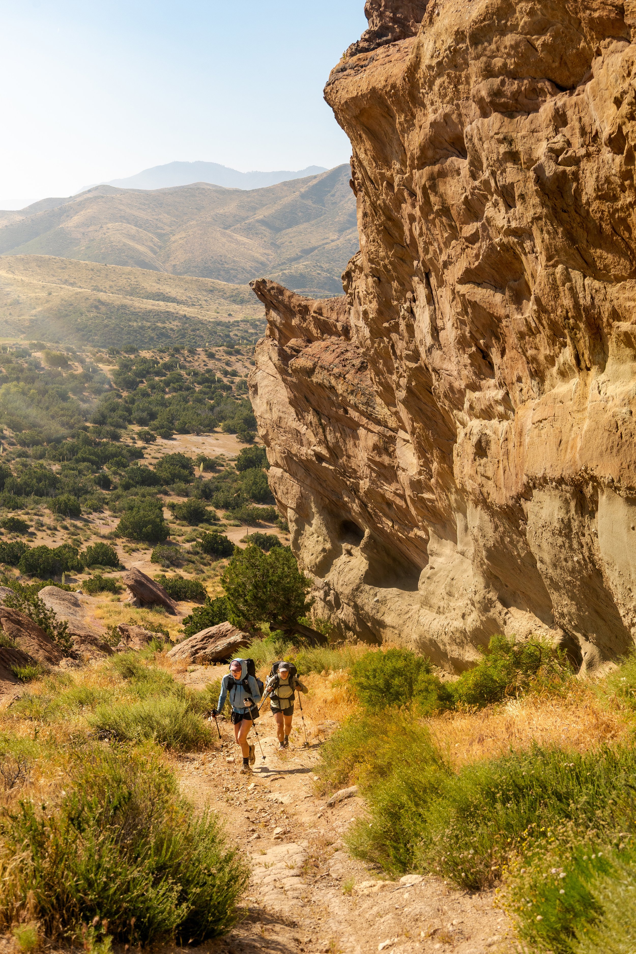 Two hikers, a man and a woman, walking on a dirt trail in a desert landscape with large rock formations on the right and mountains in the background under a clear blue sky.
