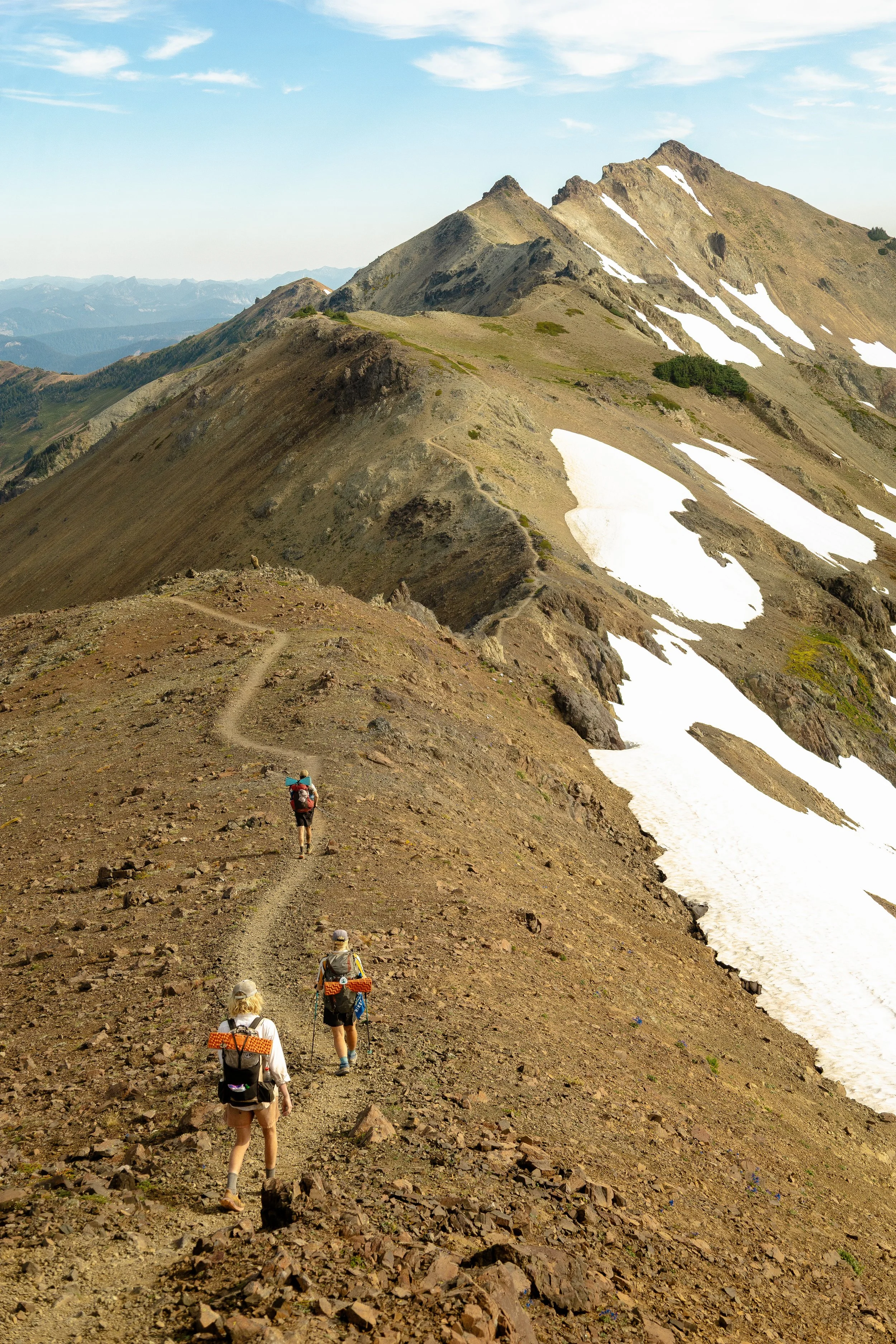 Four hikers with backpacks walking along a rocky mountain trail with mountain peaks and snow patches in the background.