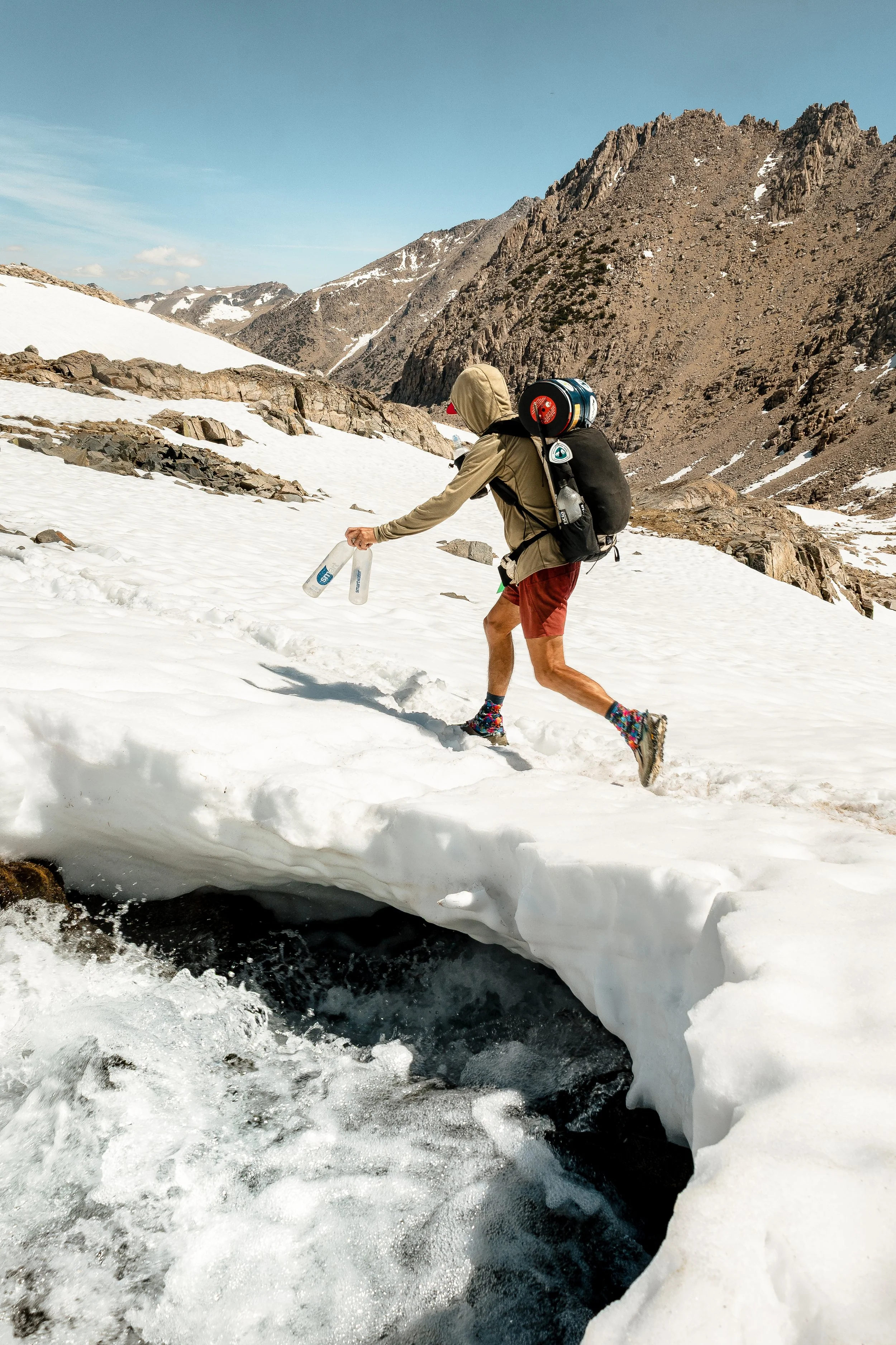 Person in hiking gear crossing a snow-covered mountain stream with mountains in the background under a clear blue sky.