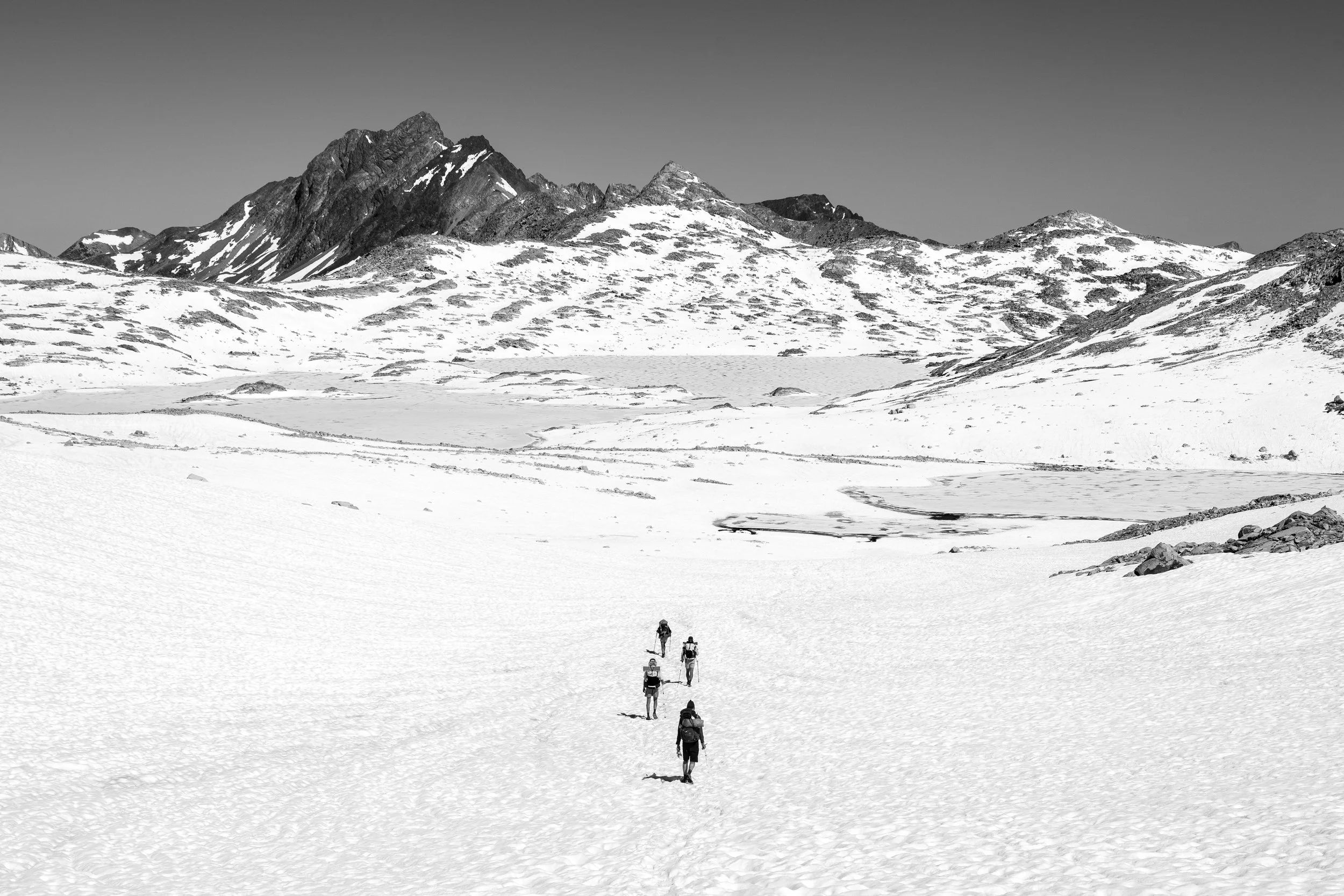 A group of hikers walking in a line across a snowy mountain landscape with rugged peaks in the background.