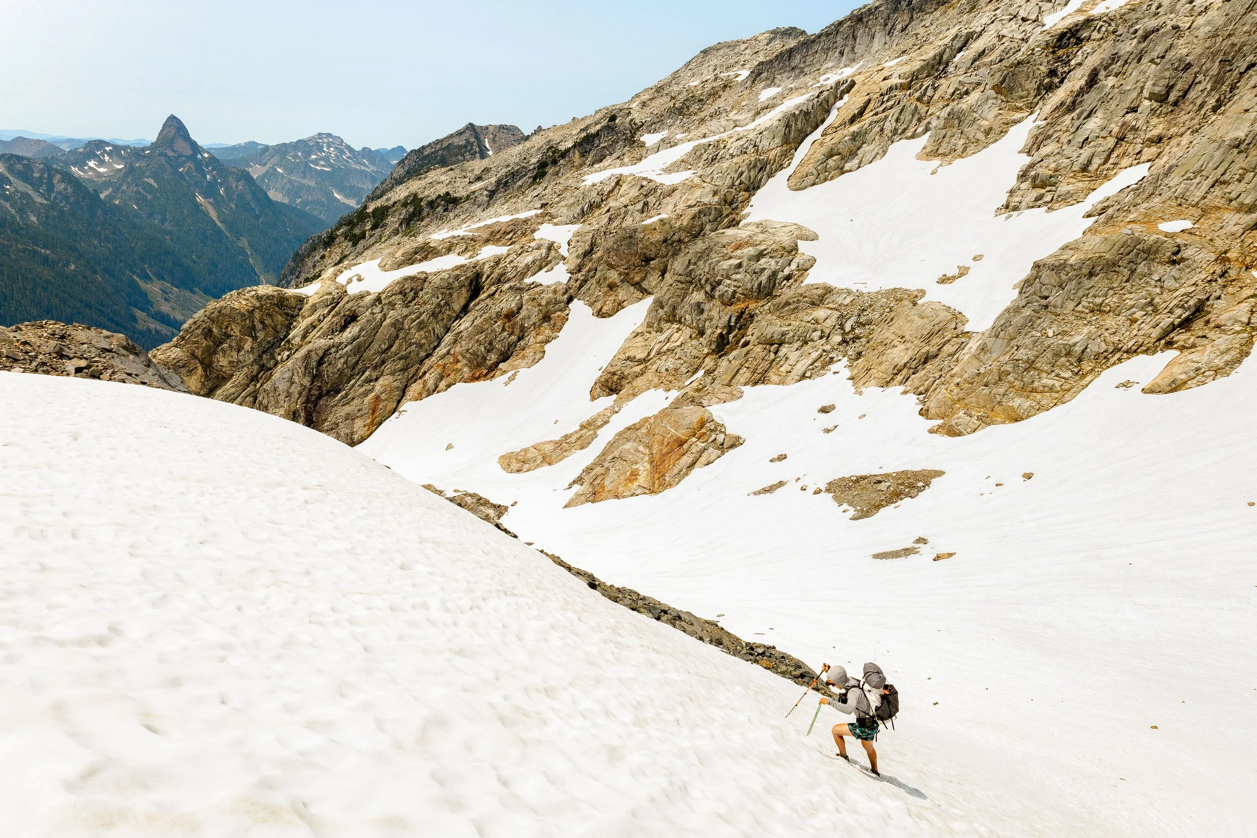 Two hikers climbing a steep snow-covered mountain slope, with rocky mountain peaks in the background.