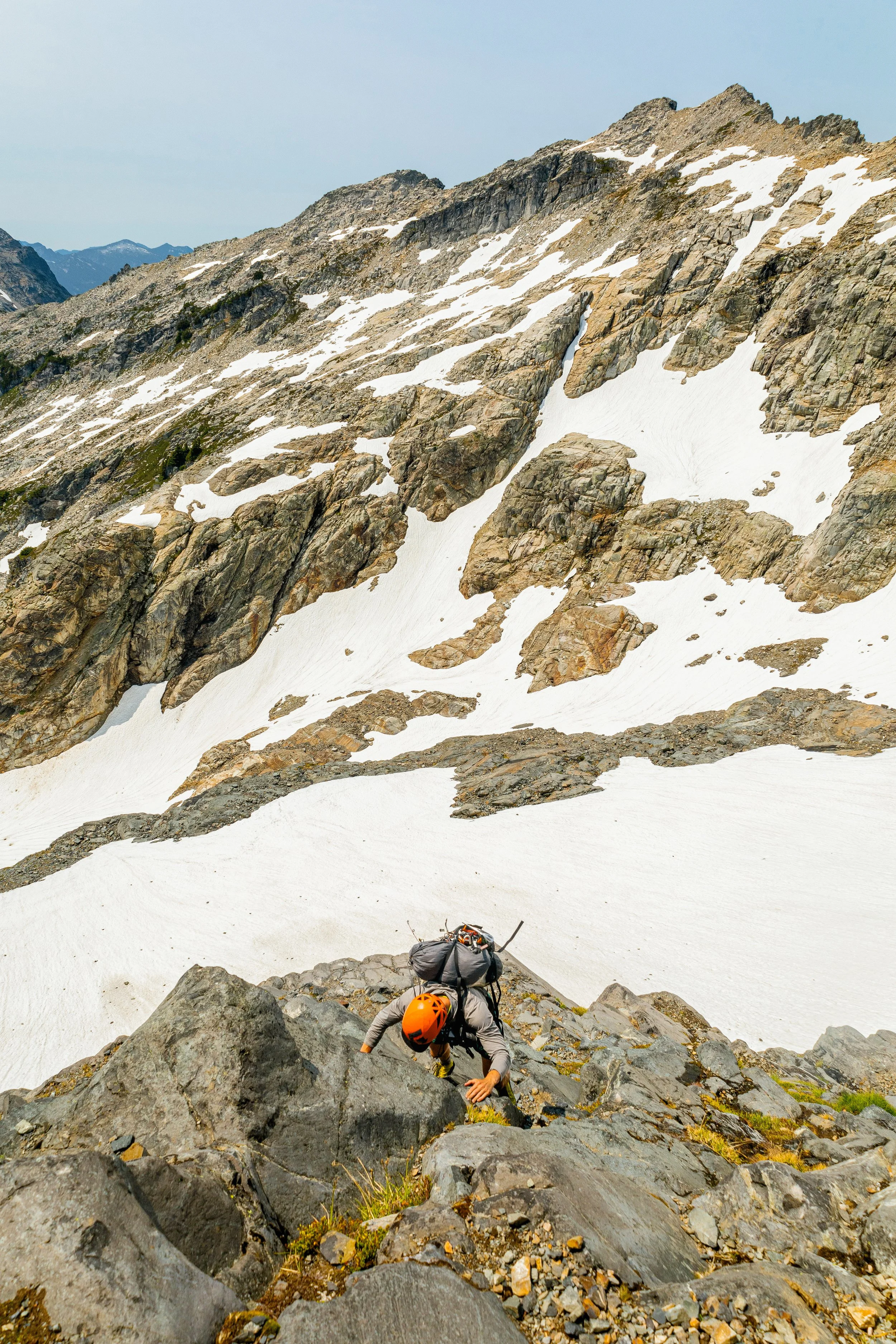 Mountain climber ascending a steep rocky slope with snow patches, wearing an orange helmet and carrying a backpack, in a rugged mountain landscape.