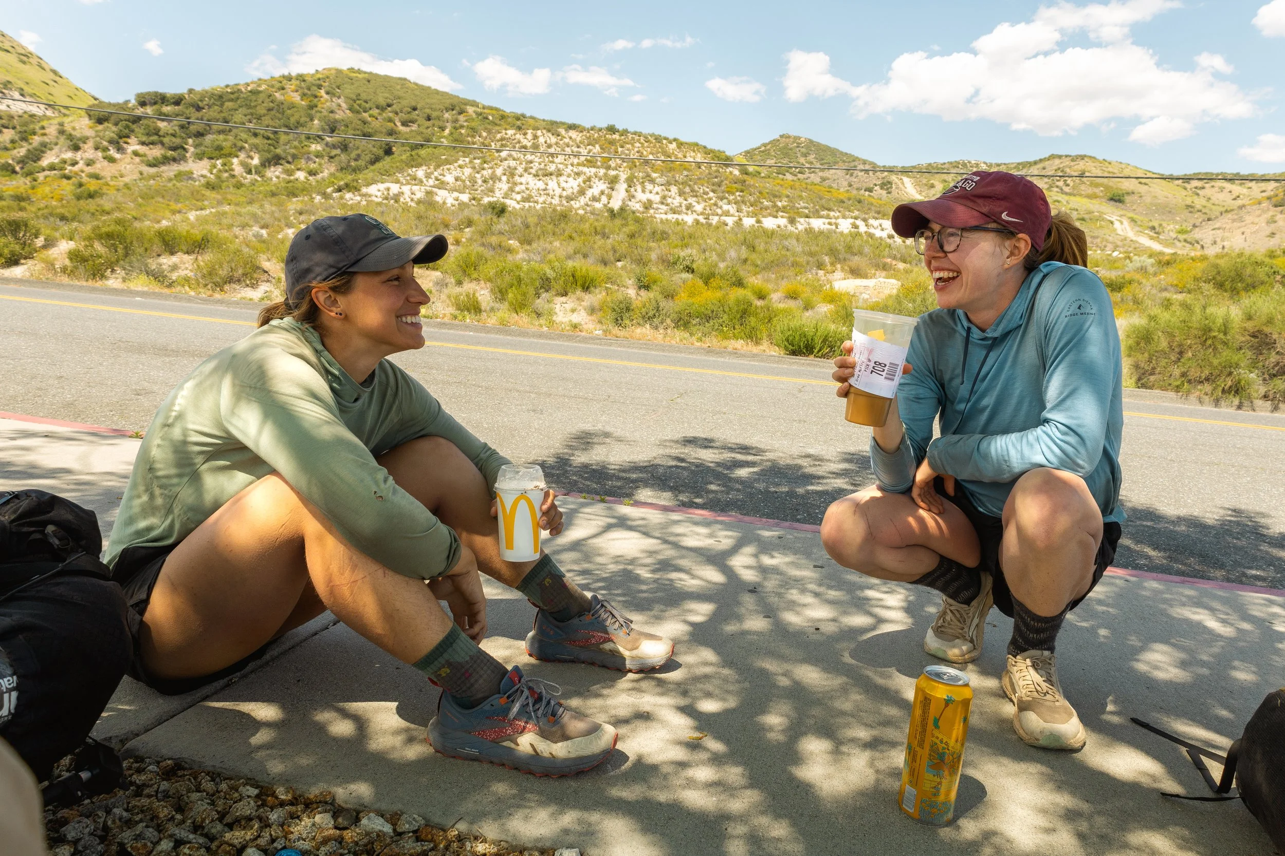 Two women resting by the roadside, sitting on the sidewalk with mountain scenery in the background, enjoying drinks and smiling at each other.